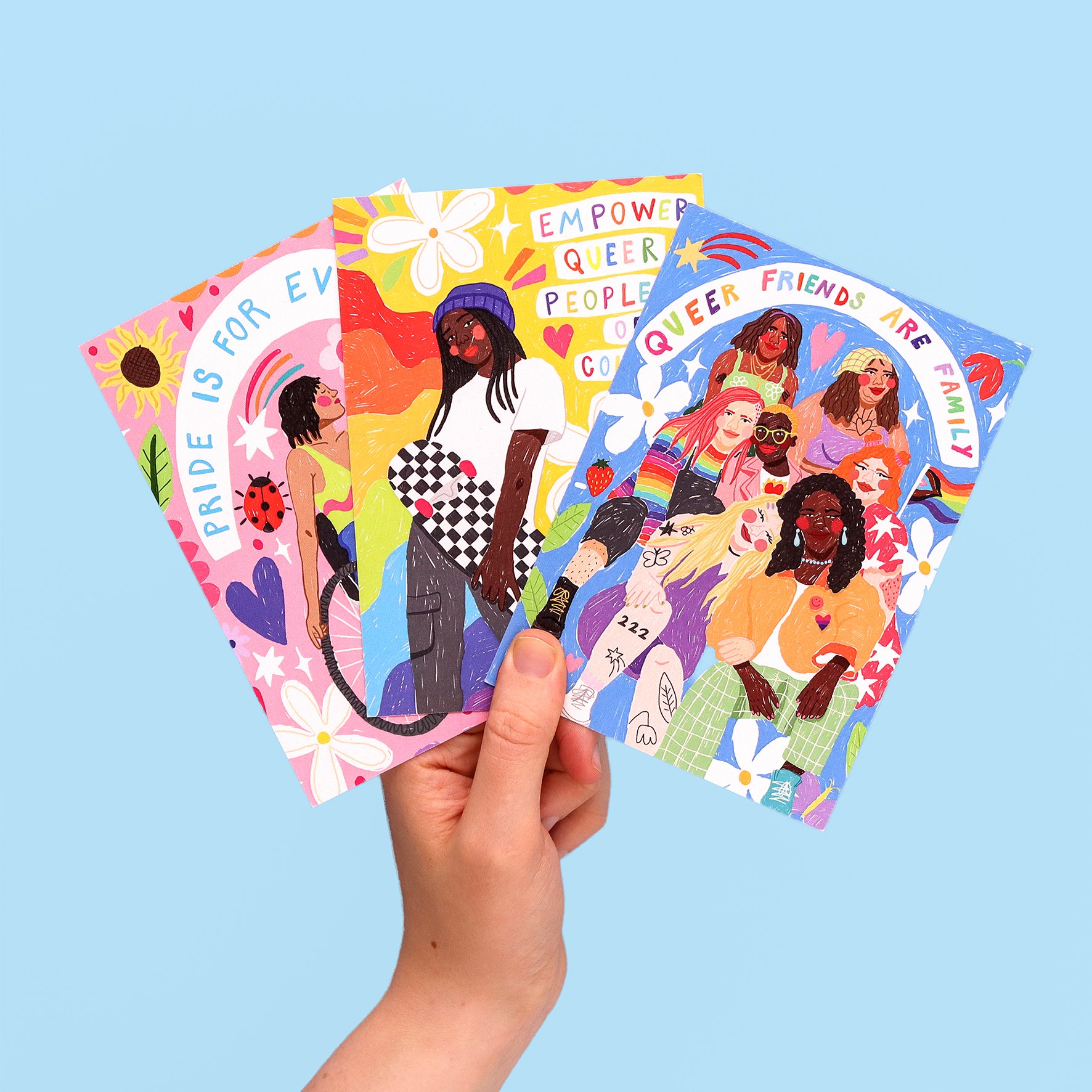 Hand holding three colorful postcards with illustrations of diverse women and empowering messages about LGBTQ+ pride and community, against a light blue background.