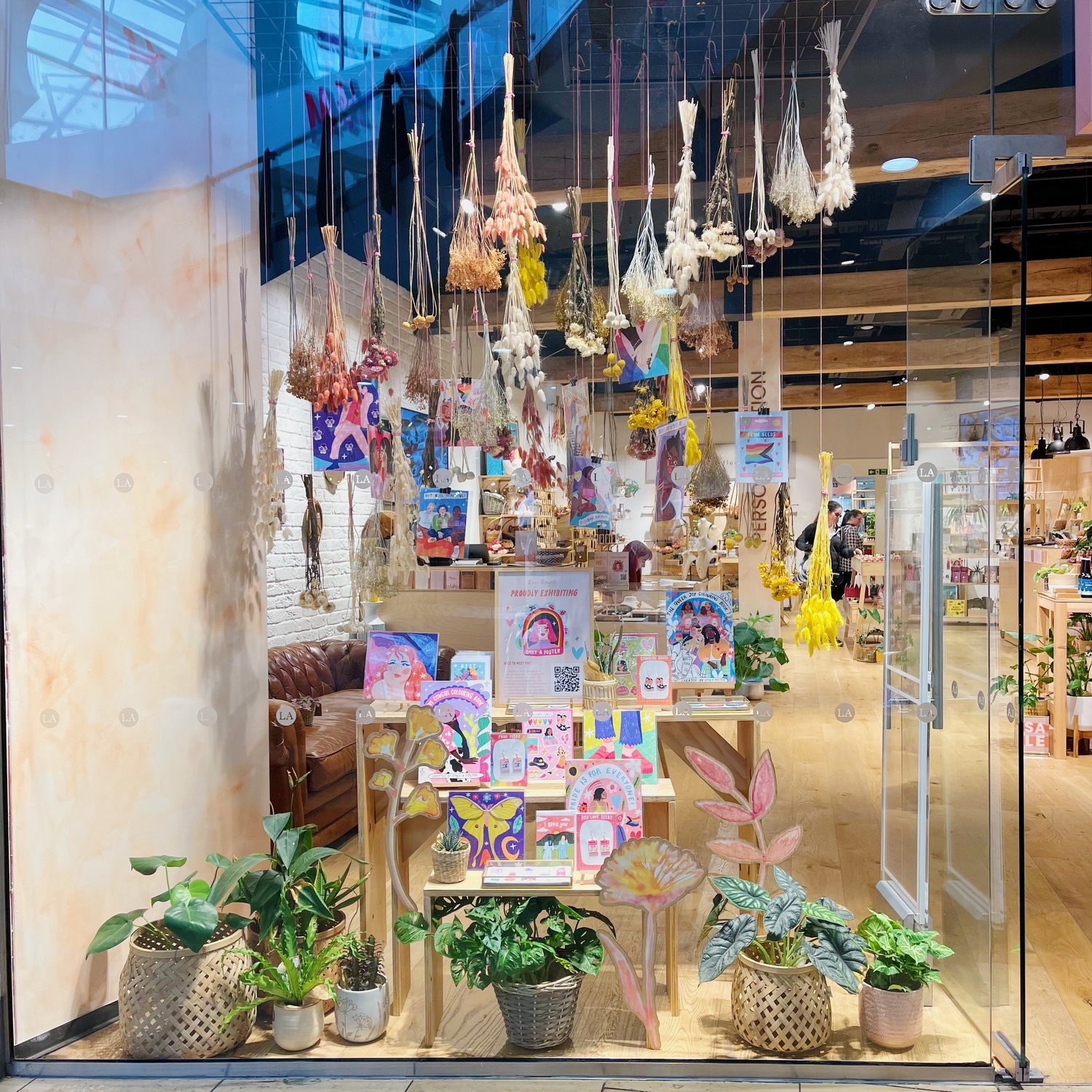 Display window of a store decorated with hanging dried flowers, indoor plants in baskets, colorful illustrated greeting cards, and artwork on the table. Inside, there are shelves with various items, plants, and customers browsing.