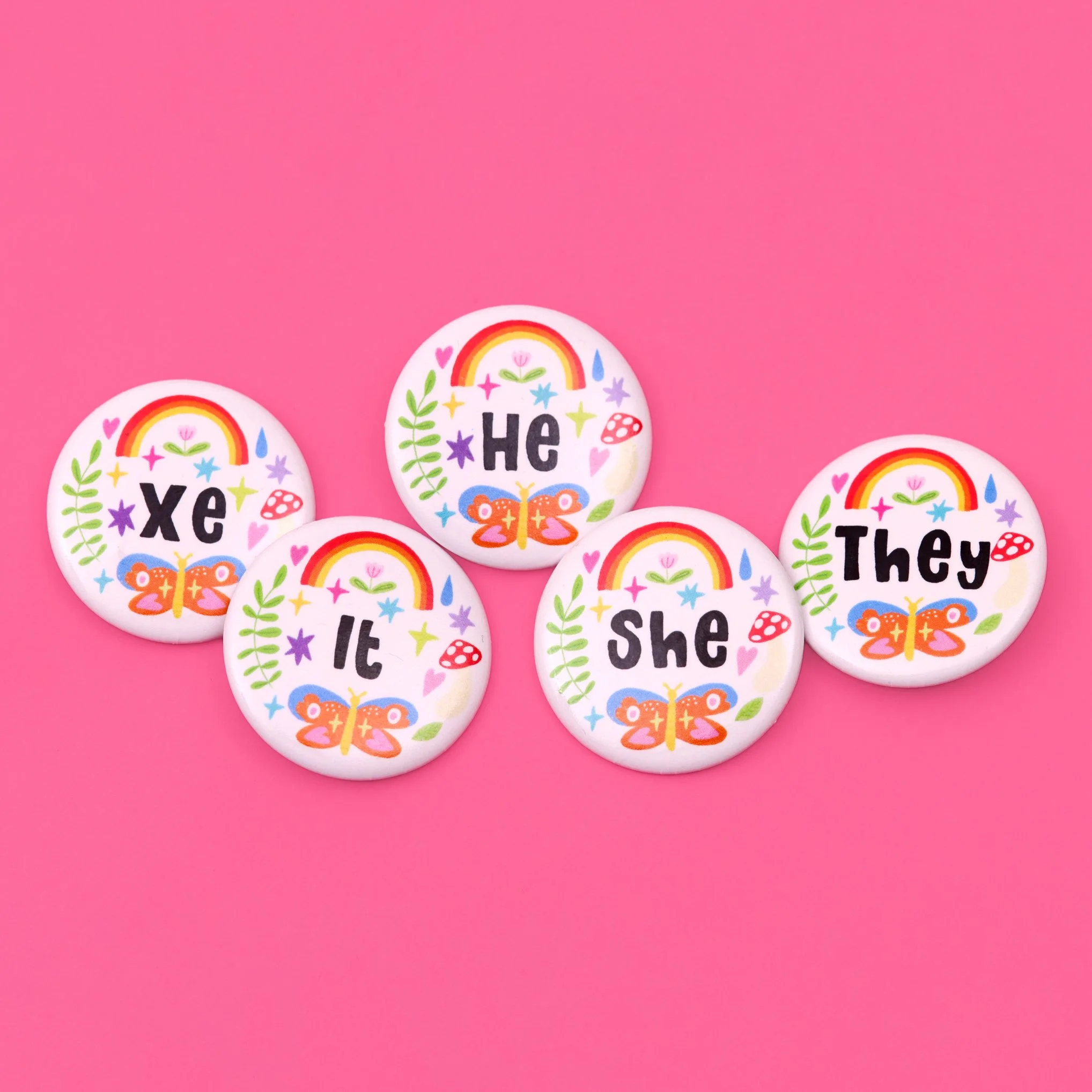 Five circular buttons with colorful designs and words 'He,' 'She,' 'They,' 'X'e,' and 'It,' decorated with rainbows, butterflies, plants, mushrooms, and stars, arranged on a pink background.