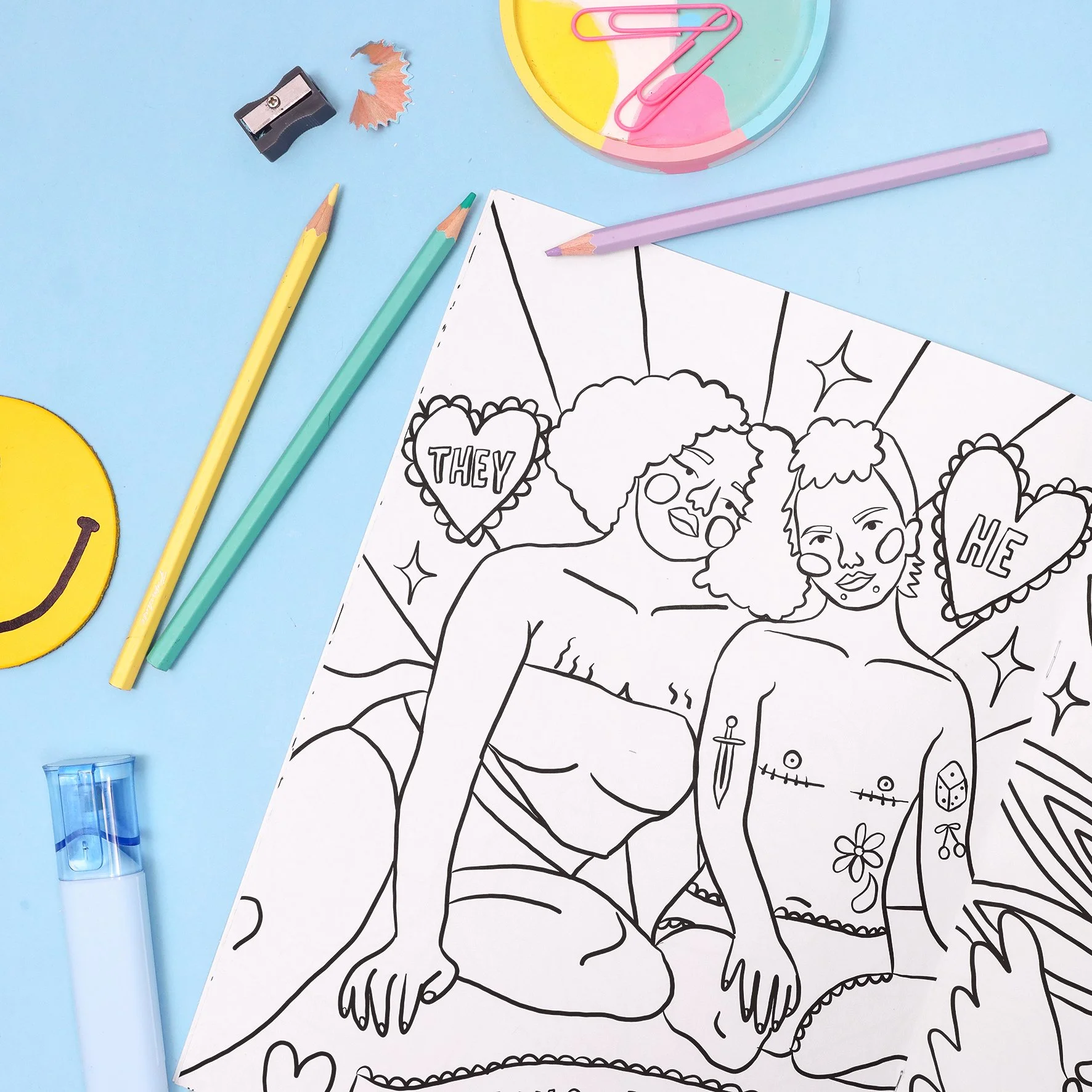 Outline drawing of two women with tattoos, some hearts, and decorative elements on a white paper, surrounded by colored pencils, a sharpener, a clip, a smiley face sticker, and a pastel-colored paper plate with pink paper clips.