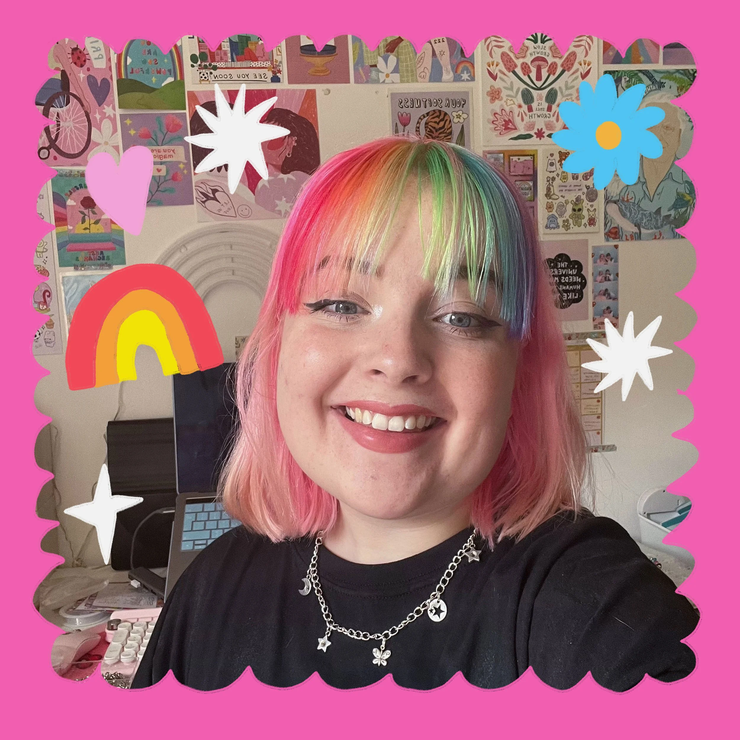 A smiling woman with short pink hair and rainbow-colored bangs, wearing a black shirt and layered silver necklaces with star and butterfly charms. The photo has a pink scalloped border and colorful doodles including hearts, stars, a rainbow, and flowers. The background features a wall decorated with various colorful posters and artwork.