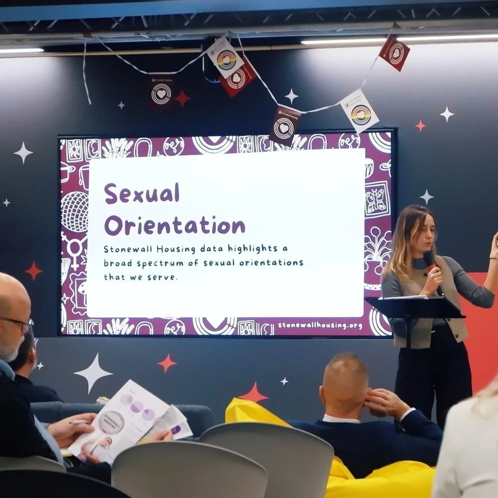 A woman presenting at a conference on a stage, with a large screen behind her displaying a slide that reads "Sexual Orientation" and mentions Stonewall Housing data highlights. Audience members are seated, some reading brochures related to the event.
