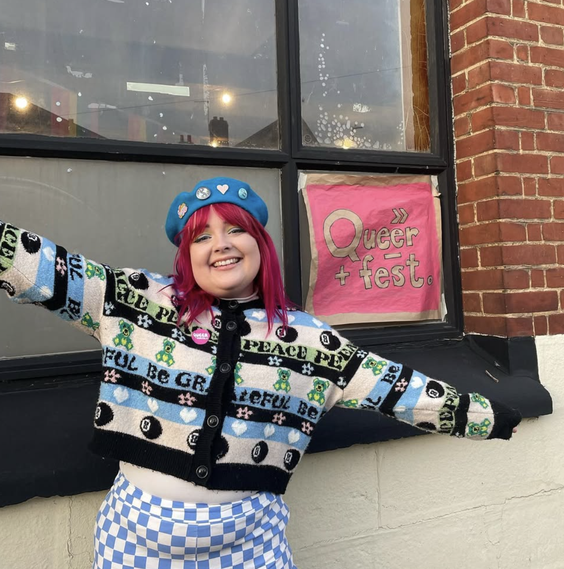 A smiling woman with pink hair wearing a blue hat with pins and a multicolored sweater, standing outside a building with a sign that says Queer + Fest.