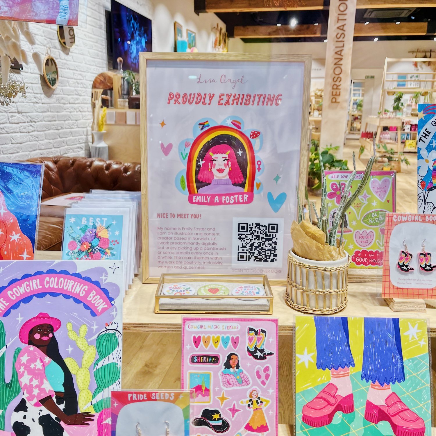 Display of colorful booklets, stickers, and artwork on a table inside a store. A framed sign announces an exhibition by Emily A Foster, featuring a cartoon portrait of a girl with pink hair. Various art pieces with vibrant designs and illustrations a