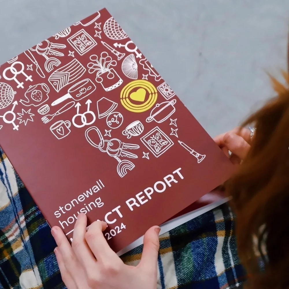 Person holding a red report booklet titled "Stonewall Housing Impact Report 2024" with various white line icons on the cover, including rainbows, keys, cups, and hearts, while wearing a plaid garment.
