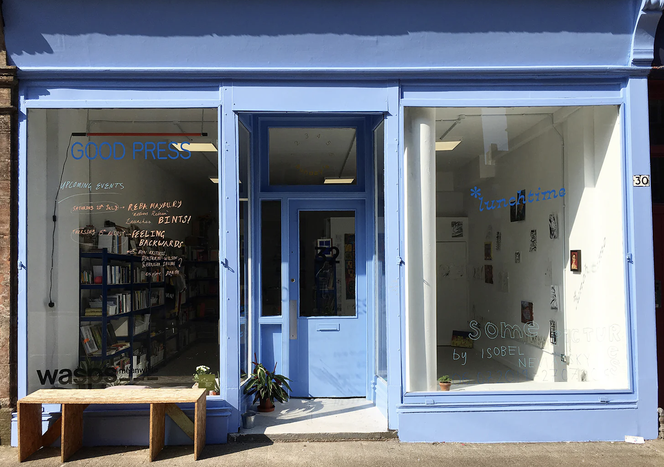 Blue storefront with large glass windows and door, inside bookshelves on the left, wall art and small plants on the right, signs with 'Good Press,' 'Lunchtime,' and handwritten event details.