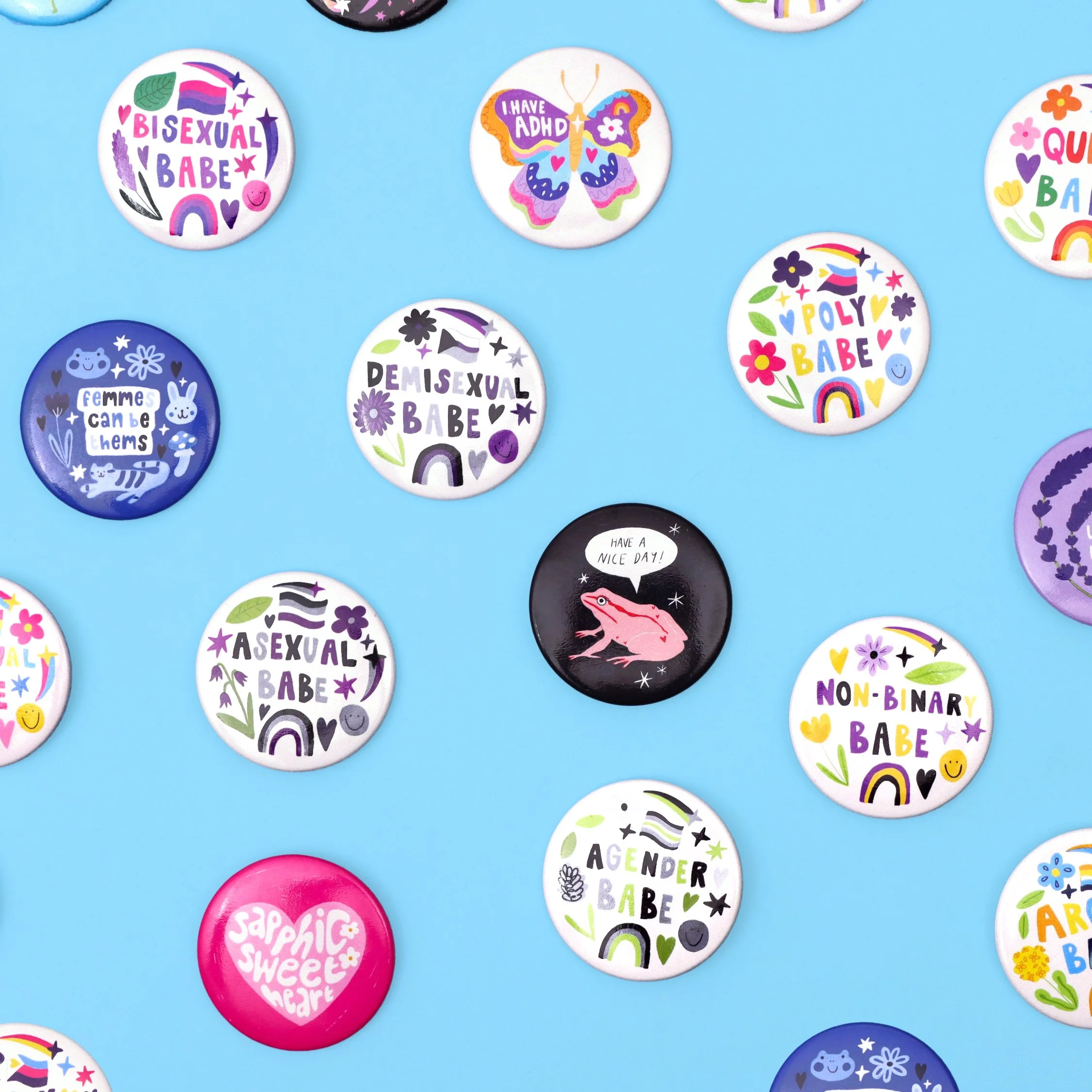 Collection of colorful buttons with various slogans and designs, including phrases like "Bisexual Babe", "I Have ADHD", "Demisexual Babe", "Poly Babe", and symbols such as rainbows, hearts, flowers, and animals, arranged on a blue background.