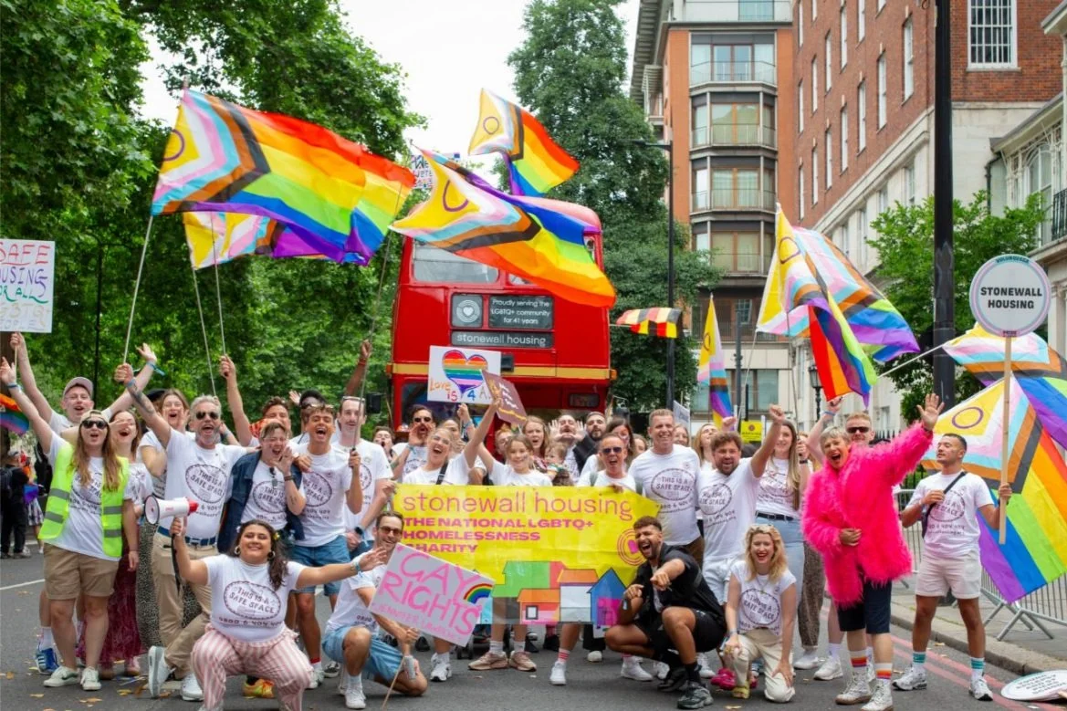 A large group of people participating in a pride parade, holding rainbow flags and banners supporting LGBTQ+ rights, with buildings and trees in the background.
