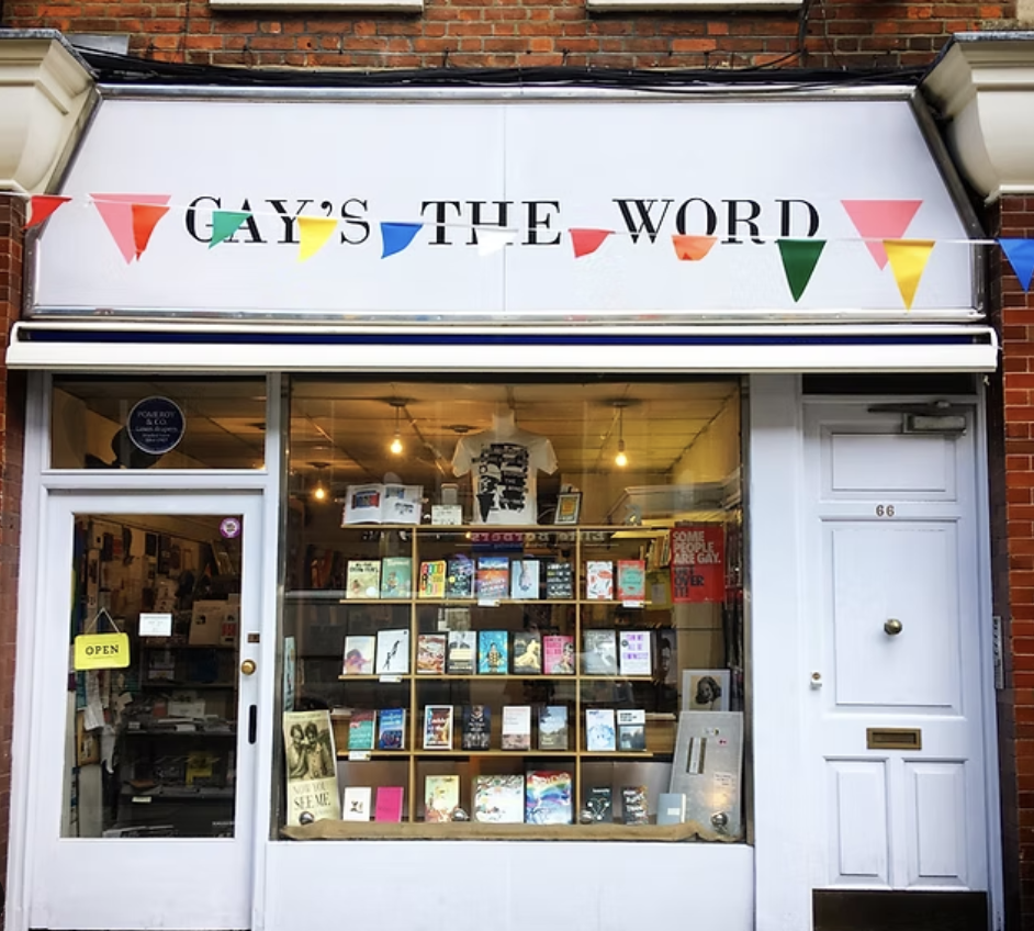 Storefront with a white sign that reads "GAYS THE WORD" decorated with colorful triangular flags. The window display showcases various books and items, with an open sign on the door.