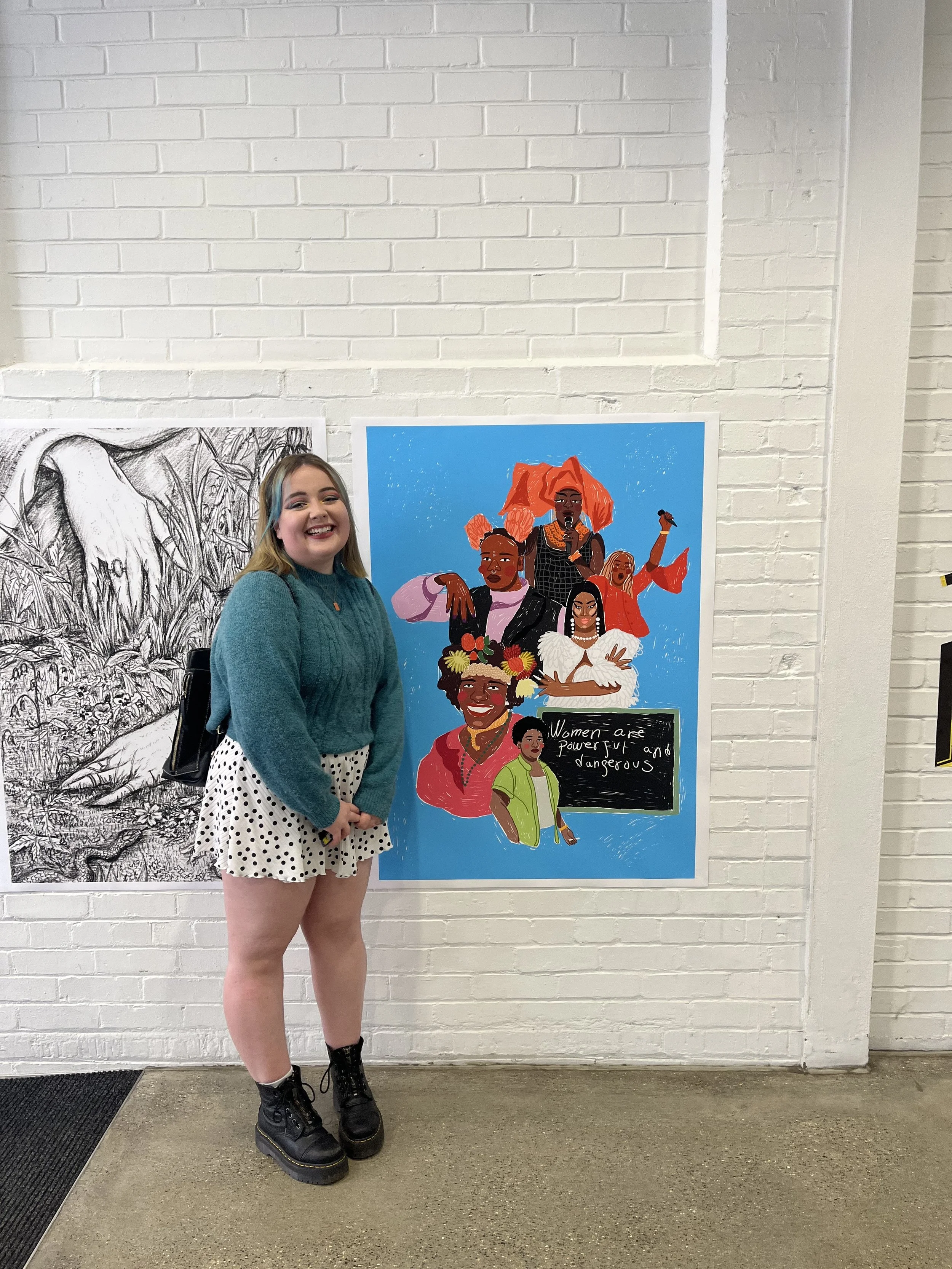 A young woman standing in front of a colorful artwork on a white brick wall, smiling at the camera. The artwork features diverse women with the quote 'Women are powerful and dangerous' on a blackboard.