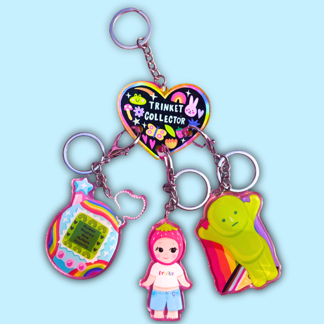 Colorful keychain with three charms: one shaped like a pink strawberry with a smiling girl, one shaped like a yellow green figure with a rainbow background, and one like a digital screen with rainbow design and the text "Queer Rights are Human Rights". The keychain is attached to a heart-shaped tag that reads "Trinket Collector" with colorful cartoon characters and designs.