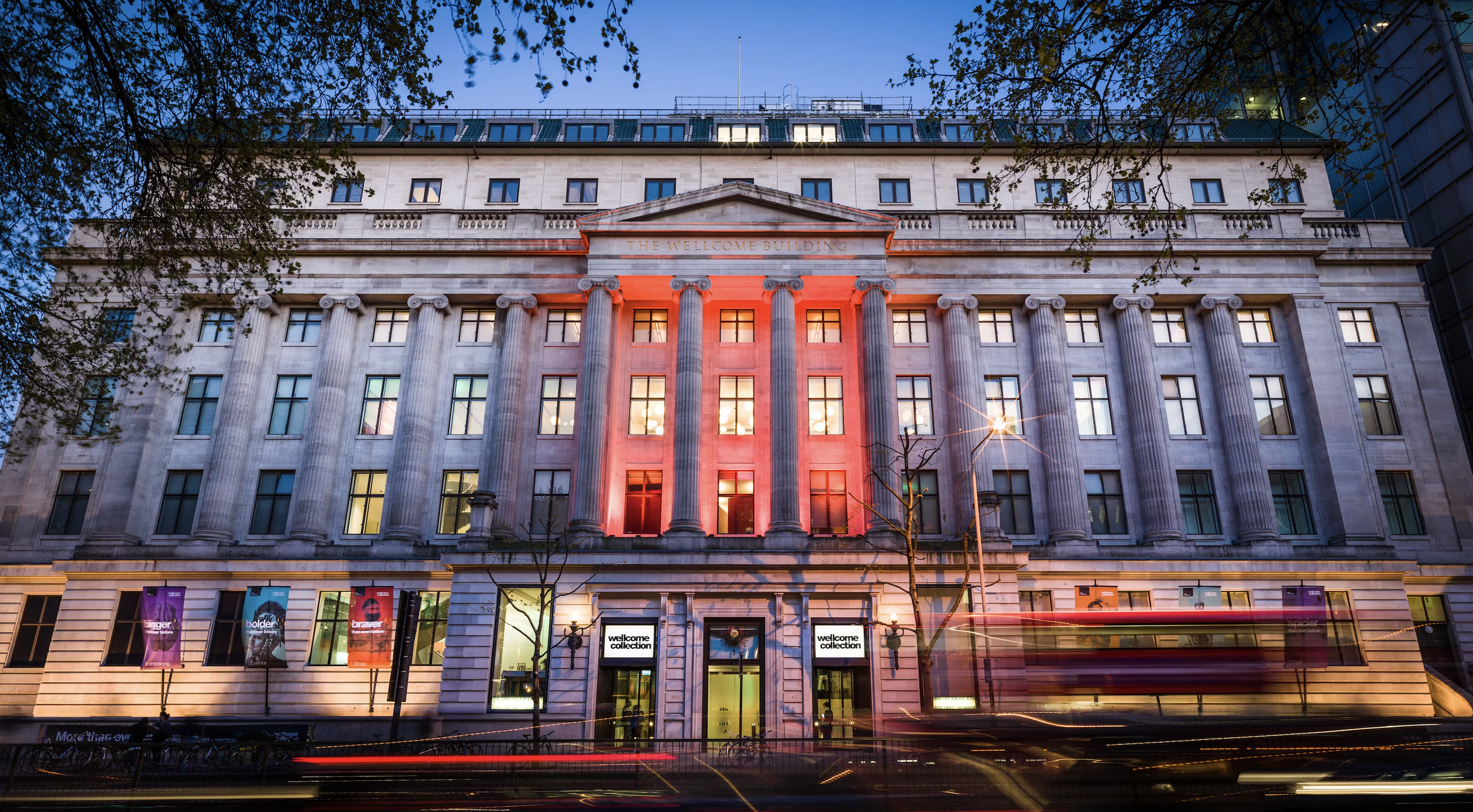 Front view of a historic building with large columns, illuminated in red lights, with a city street in front showing blurred lights from moving vehicles, at dusk.