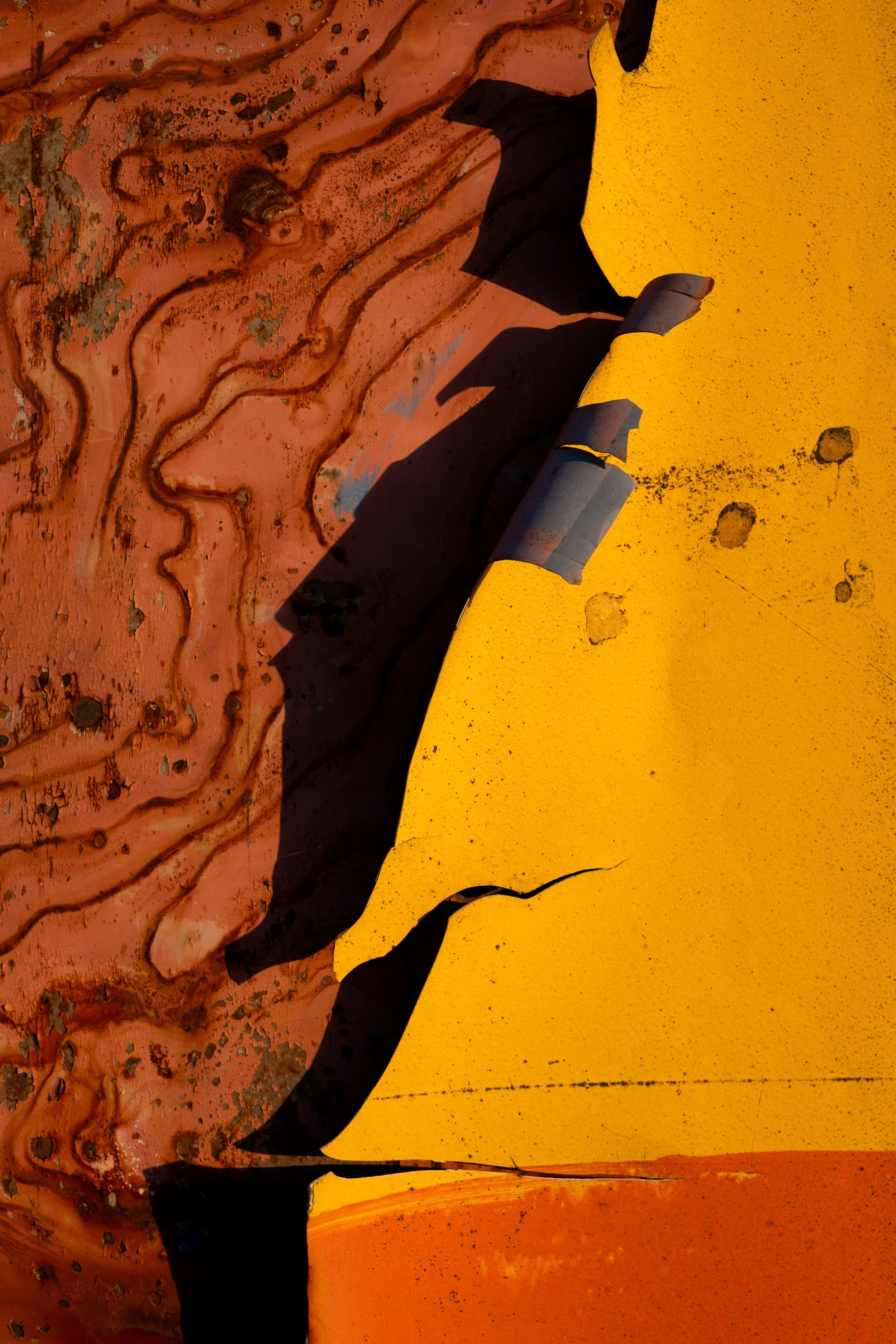 Turning a new chapter: Close-up of peeling yellow paint on a metal surface, casting a shadow, with an adjacent rusted, textured surface.
