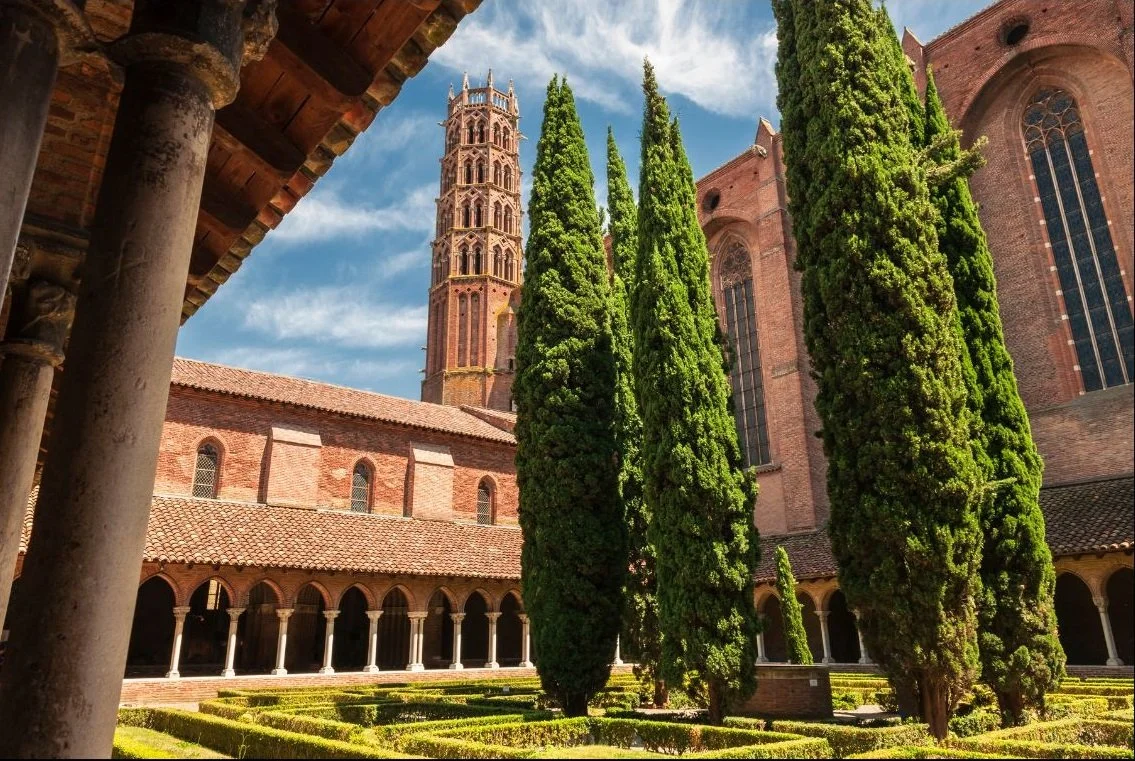 Jardin avec des arbres en colonnes, bâtiments en briques avec des fenêtres gothiques, tour d'horloge en arrière-plan, ciel bleu avec quelques nuages.