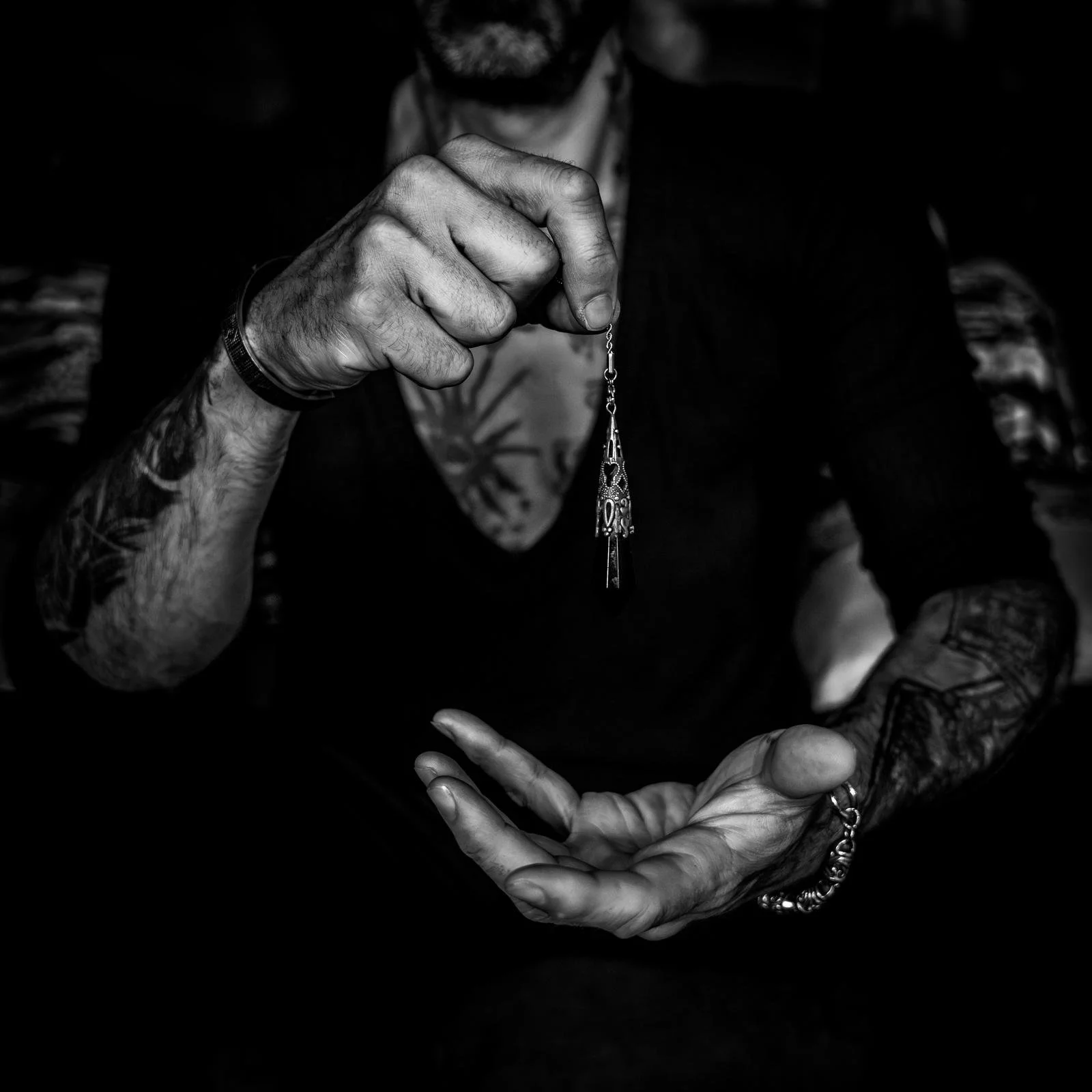 Une personne tatouée avec une barbe, portant des bracelets, tient un pendentif en métal délicat entre ses doigts. L'image est en noir et blanc, le fond est sombre.