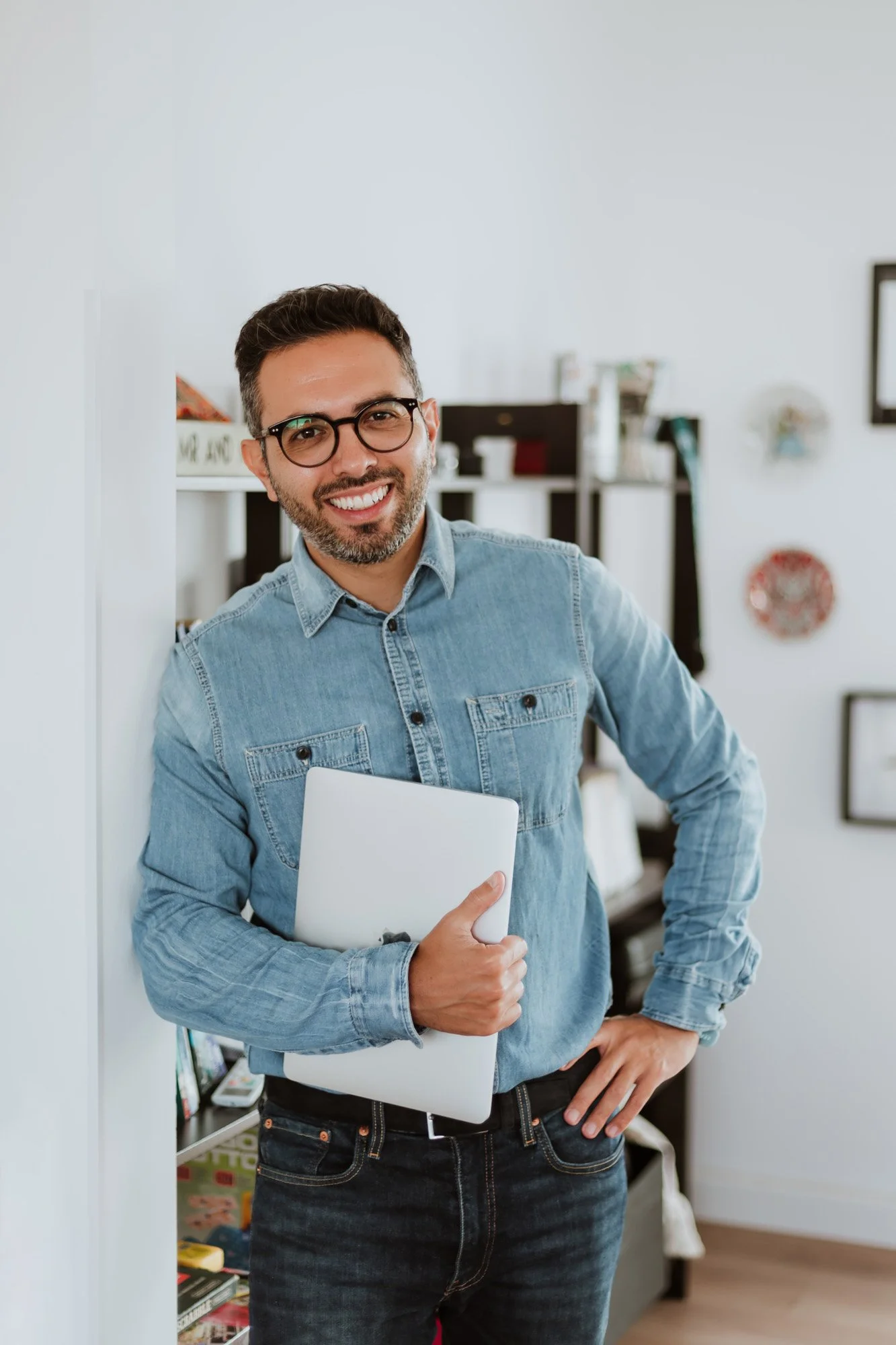 Smiling man with glasses holding a closed laptop, standing in a room with shelves and artwork on the wall.