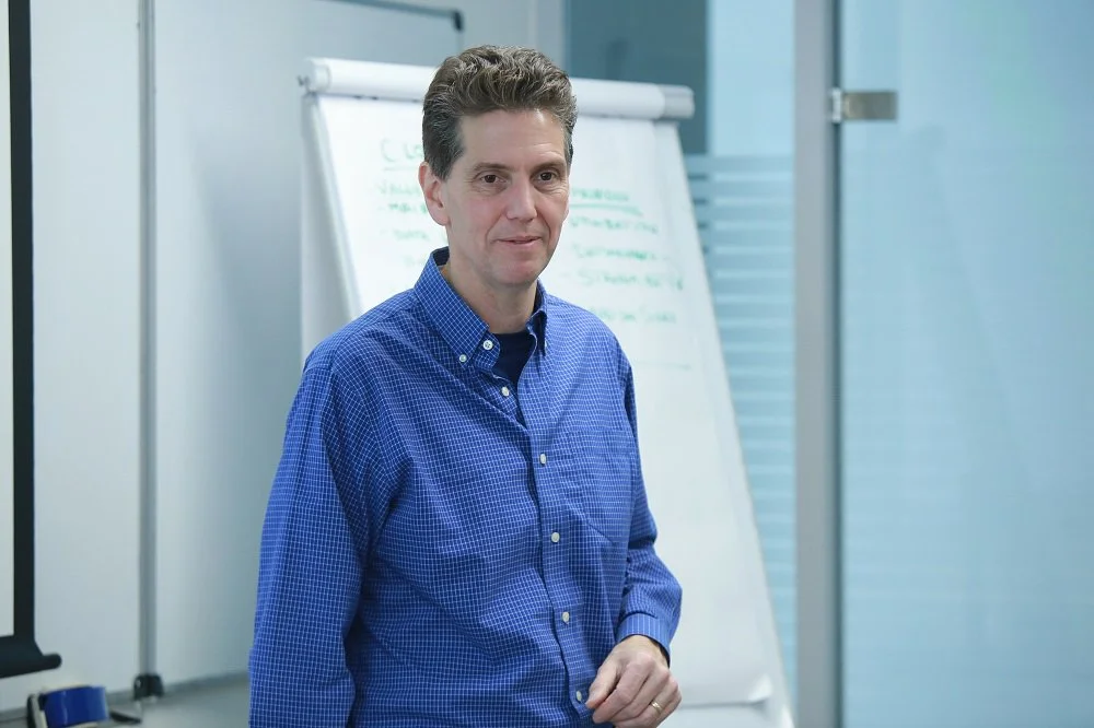 Man in blue checkered shirt standing in front of a whiteboard in a classroom or business setting.