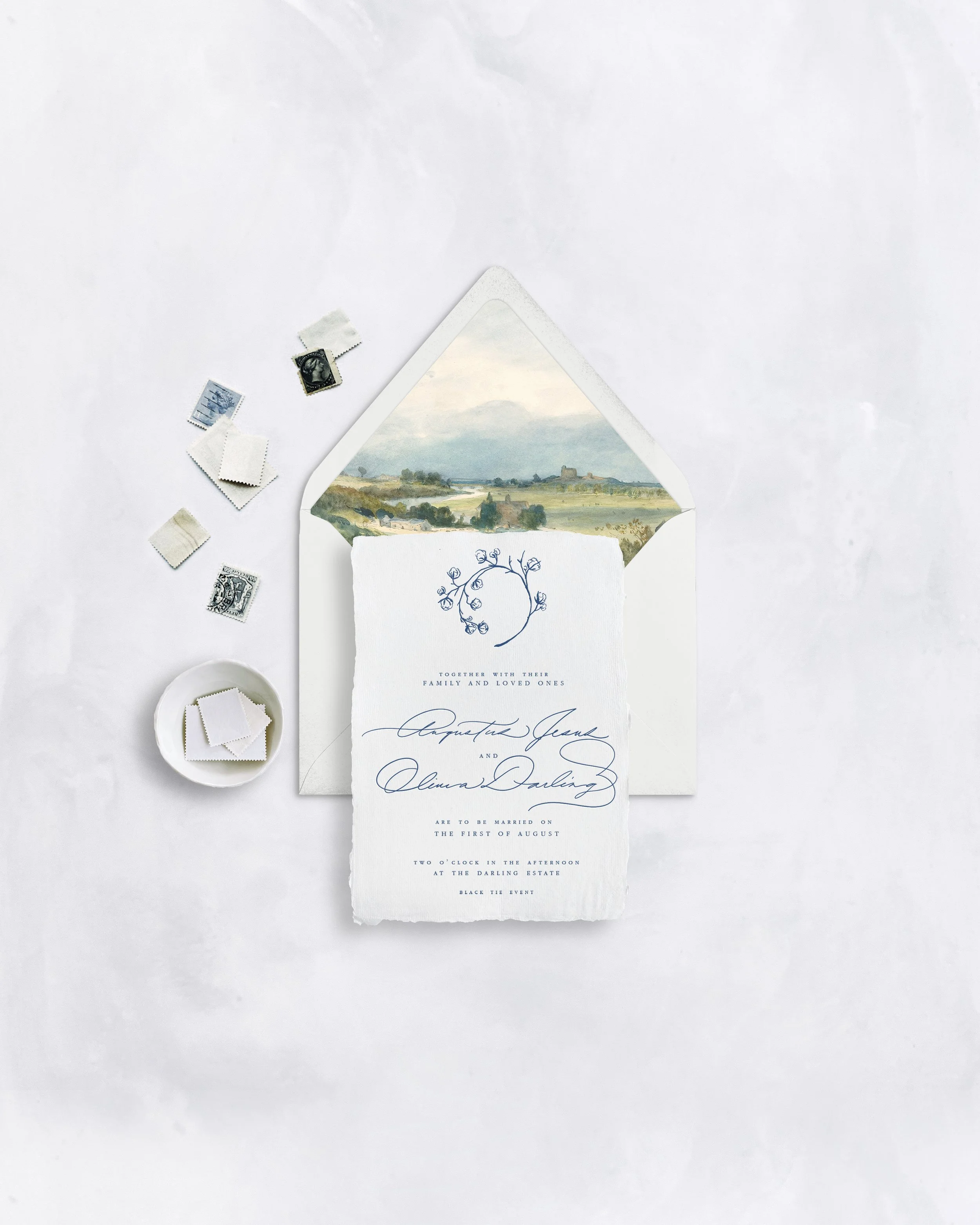 A wedding invitation with a landscape painting, a white ribbon, and a card with floral sketch, surrounded by postage stamps and a small dish of stamps on a white background.