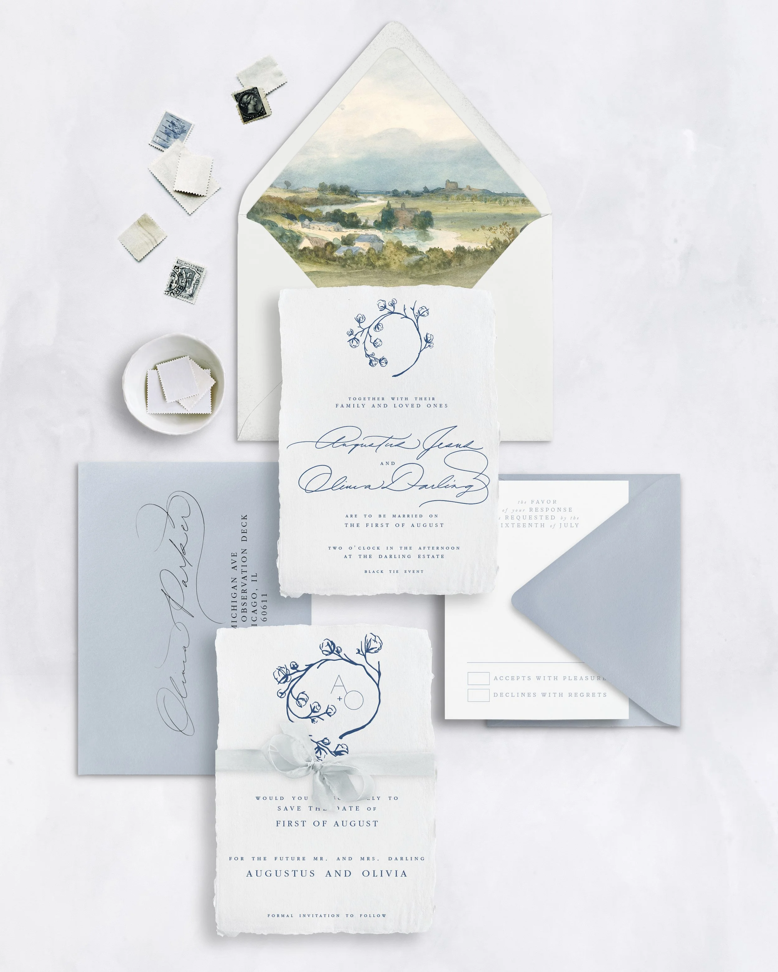 Invitation suite for a wedding, including a save-the-date card, wedding invitation, RSVP card, and envelope with address, all placed on white fabric with small stamps, a small bowl of additional stamps, and a partially opened envelope with a landscape painting inside.