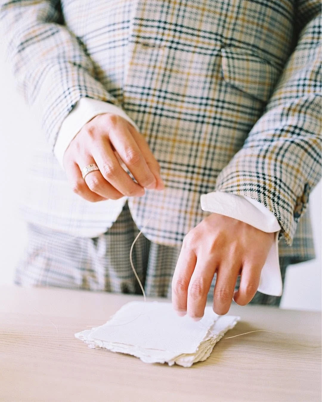 Person handling a needle and white fabric with frayed edges, wearing a checkered blazer and jewelry, in a well-lit room.
