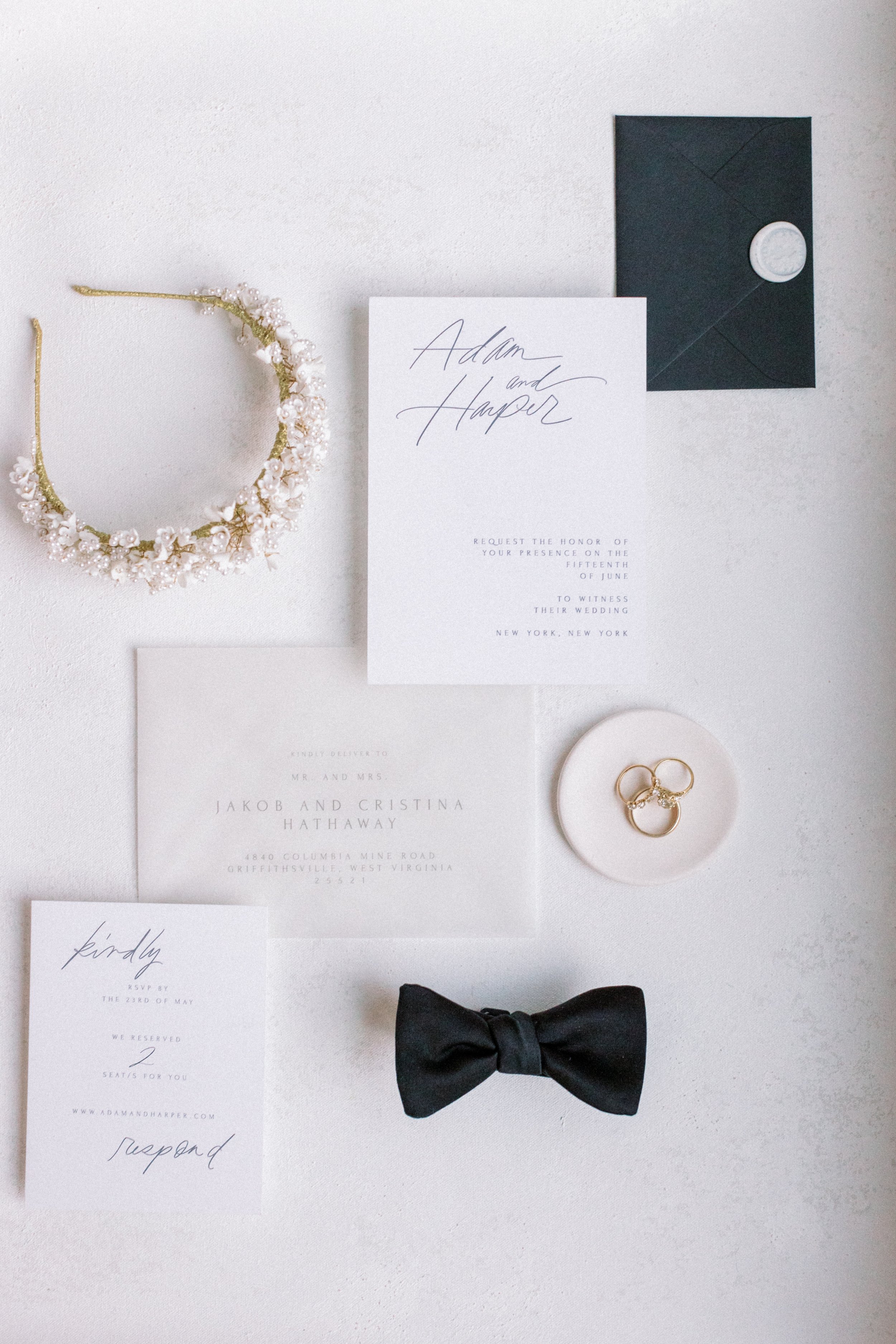 Wedding invitation suite with a black tuxedo bow tie, wedding bands on a white dish, black envelope with seal, pearl and gold tiara, and RSVP card.