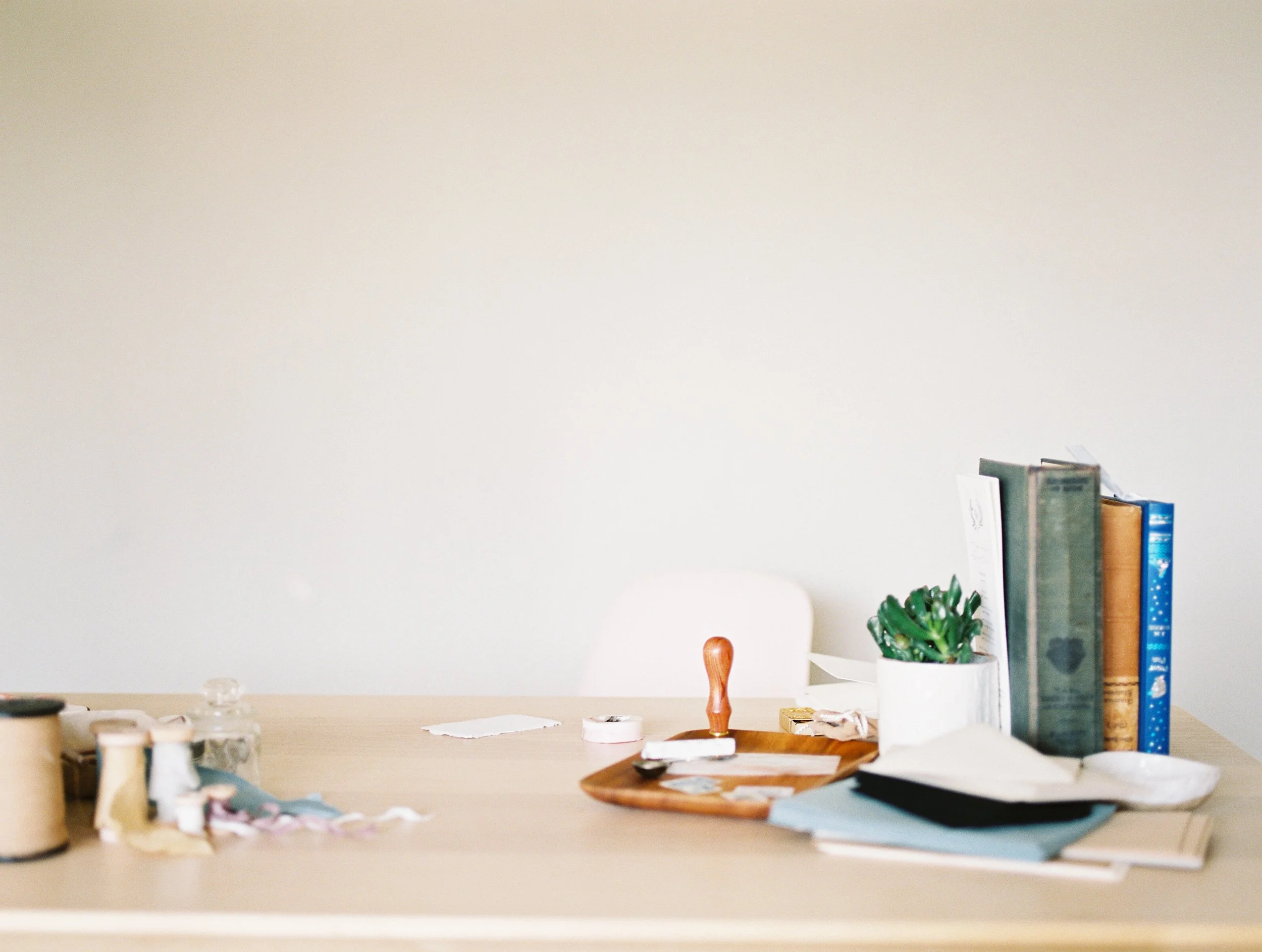 A light wooden table with various sewing supplies, a small potted succulent, stack of books, glue, scissors, and papers, against a plain white wall.