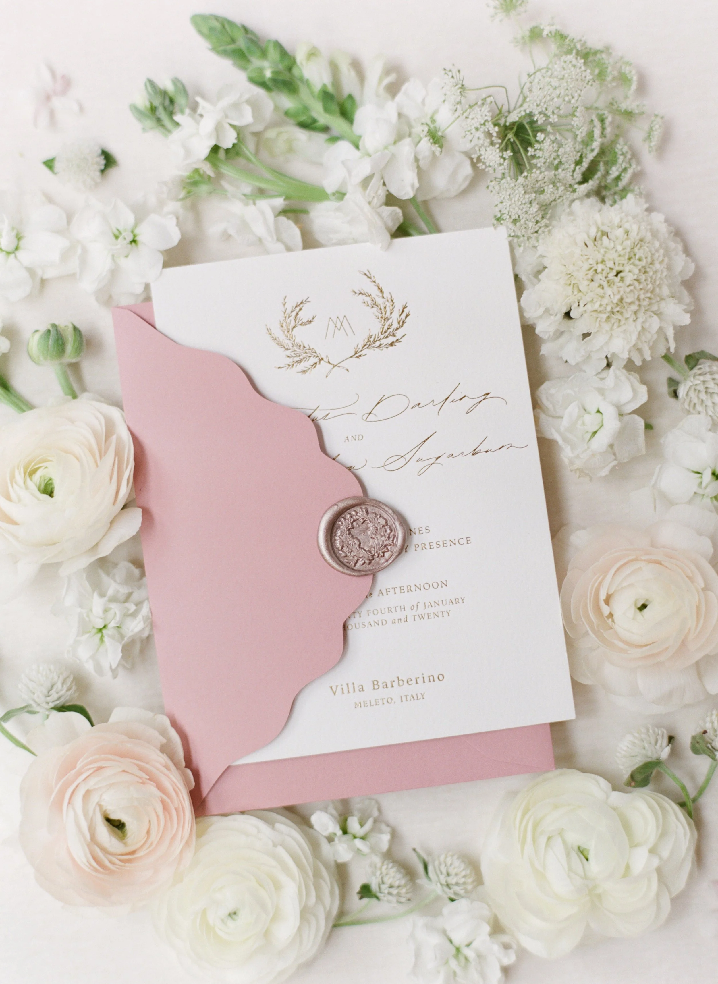Elegant wedding invitation with pink envelope and floral decorations, featuring gold script and a wax seal, surrounded by white and blush flowers.