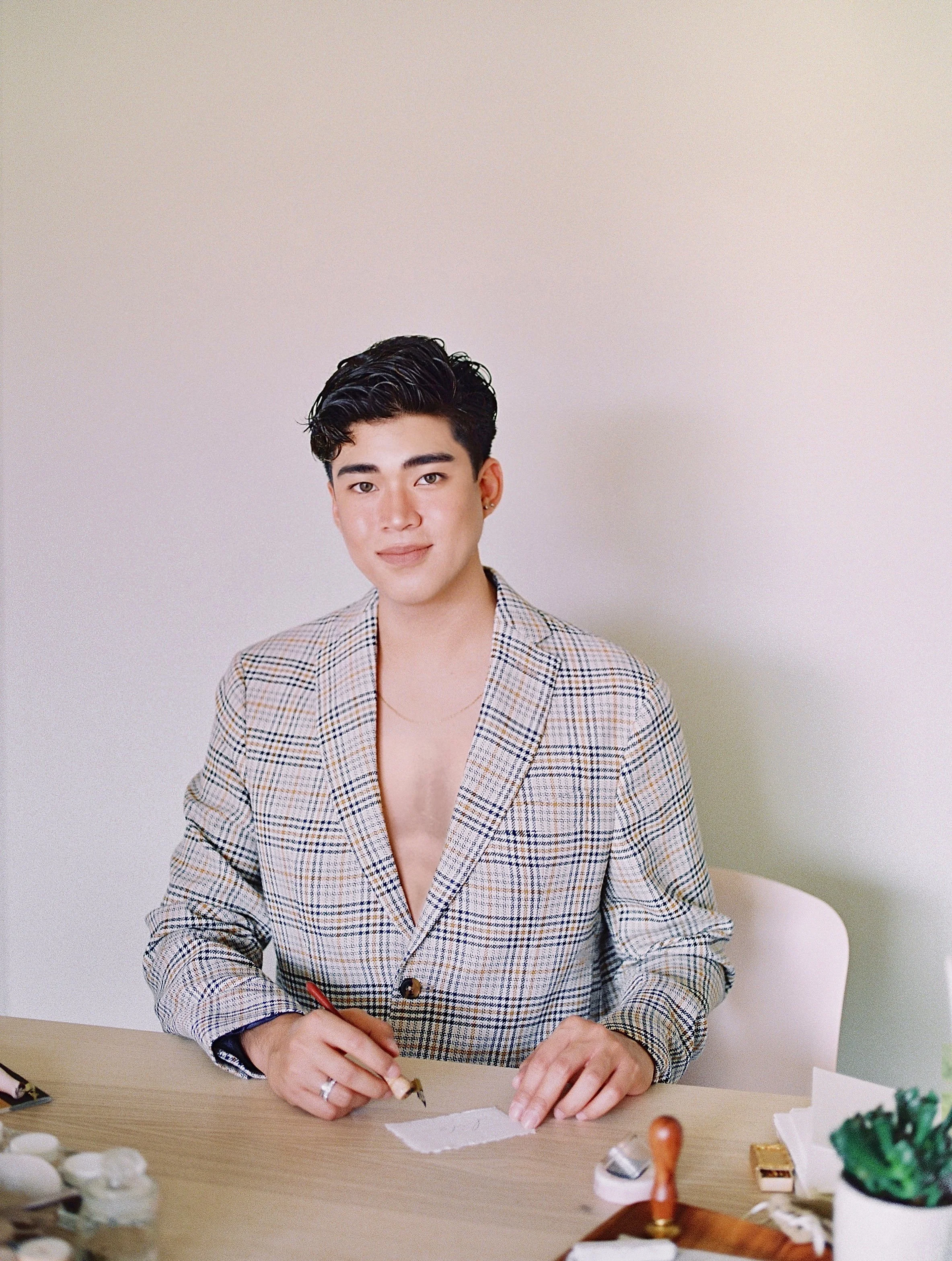 Young man with short black hair wearing a checkered blazer, seated at a table with art and craft supplies.
