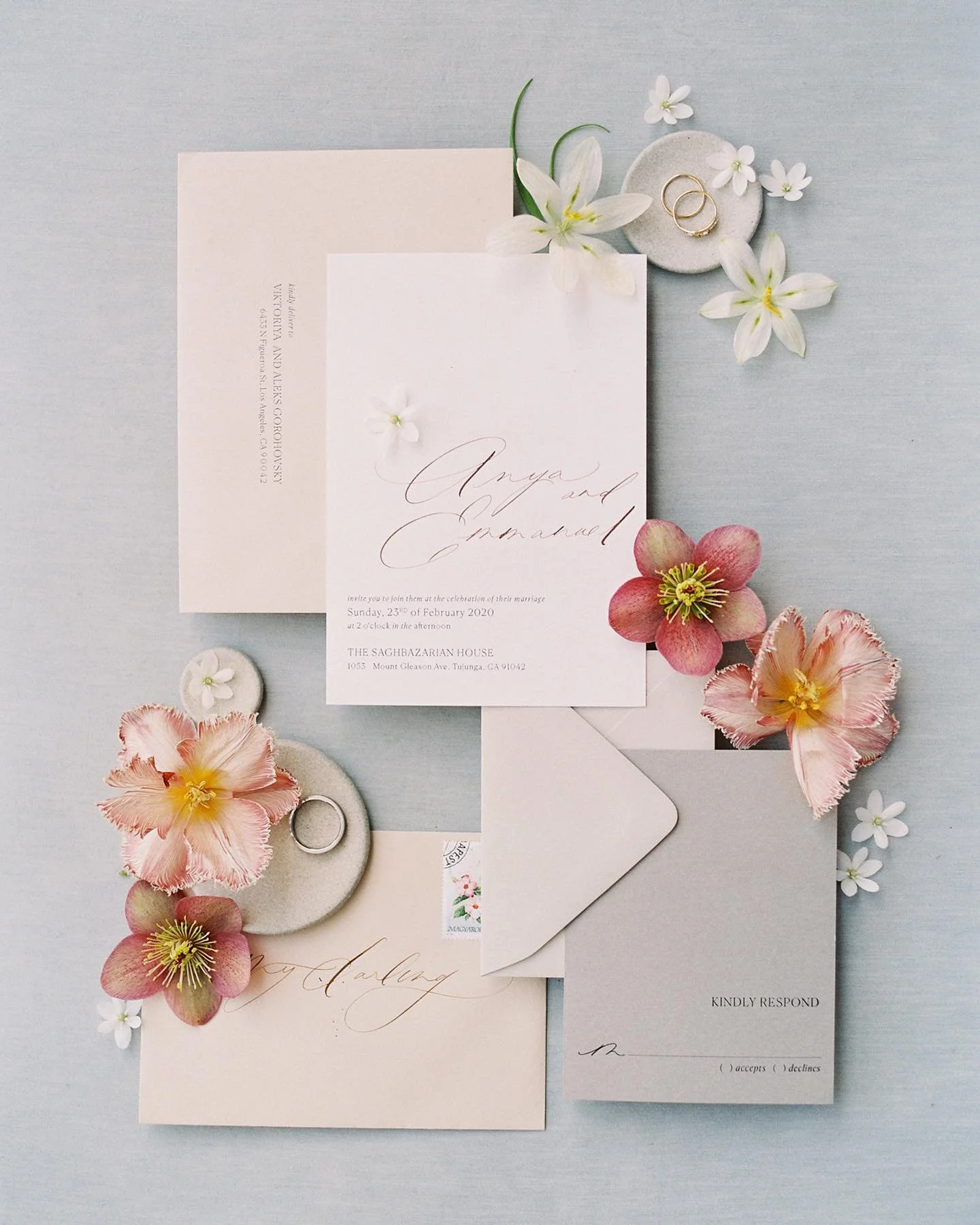 Invitation suite with wedding details, surrounded by pink and white flowers and rings on a small gray dish, against a light blue background.