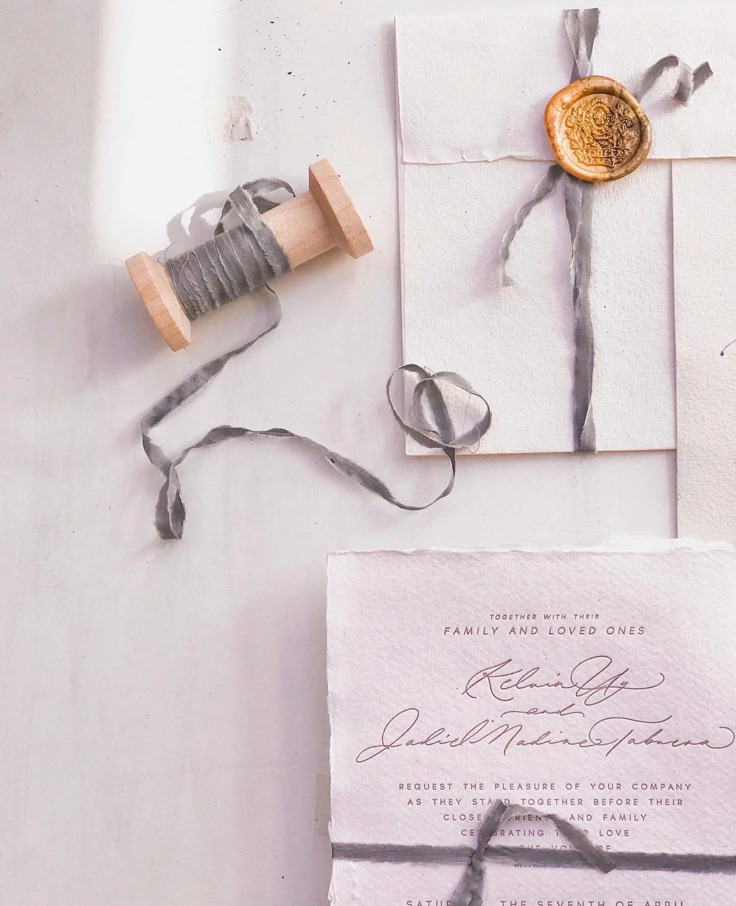 A white textured background with a spool of gray ribbon, a white card, a handwritten wedding invitation, and a wax seal stamp with gray ribbon.