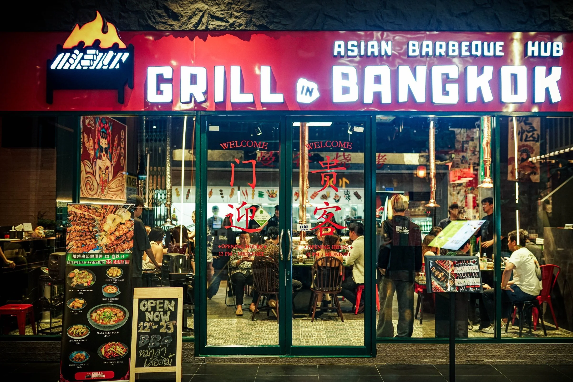 The exterior of a restaurant named Grill in Bangkok, with a large red sign overhead displaying the name and Asian barbecue concept. The glass doors have red Chinese characters and welcome signs, revealing a busy interior with diners, staff, and menu