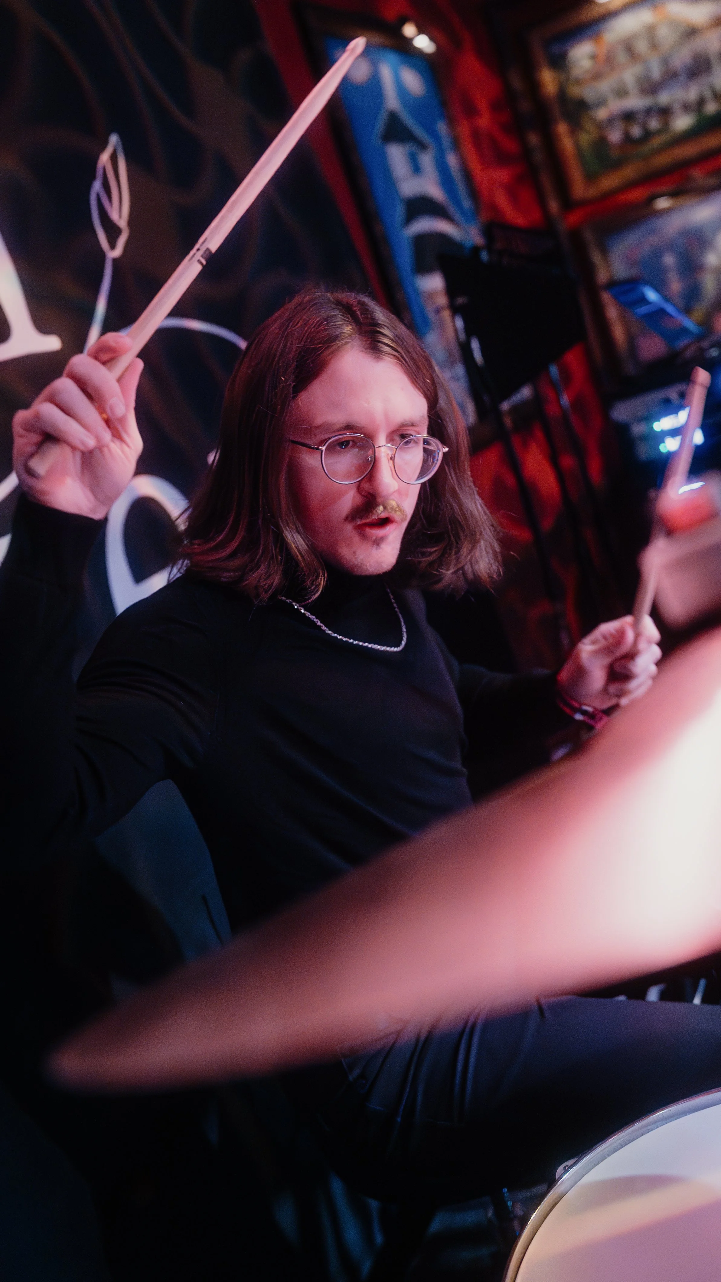 Kash Jaeger with shoulder-length hair, glasses, and a mustache, playing the drums in a dimly lit room with colorful artwork and posters on the walls.