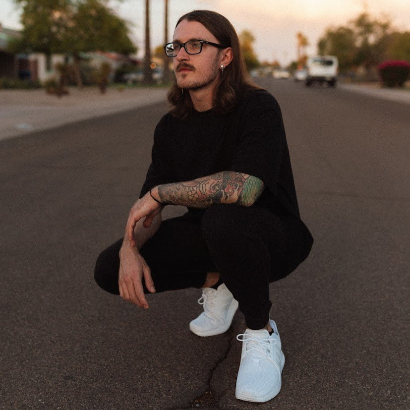 Kash Jaeger with long hair, glasses, and tattoos squatting on an empty street during sunset, wearing a black t-shirt, black pants, and white sneakers.