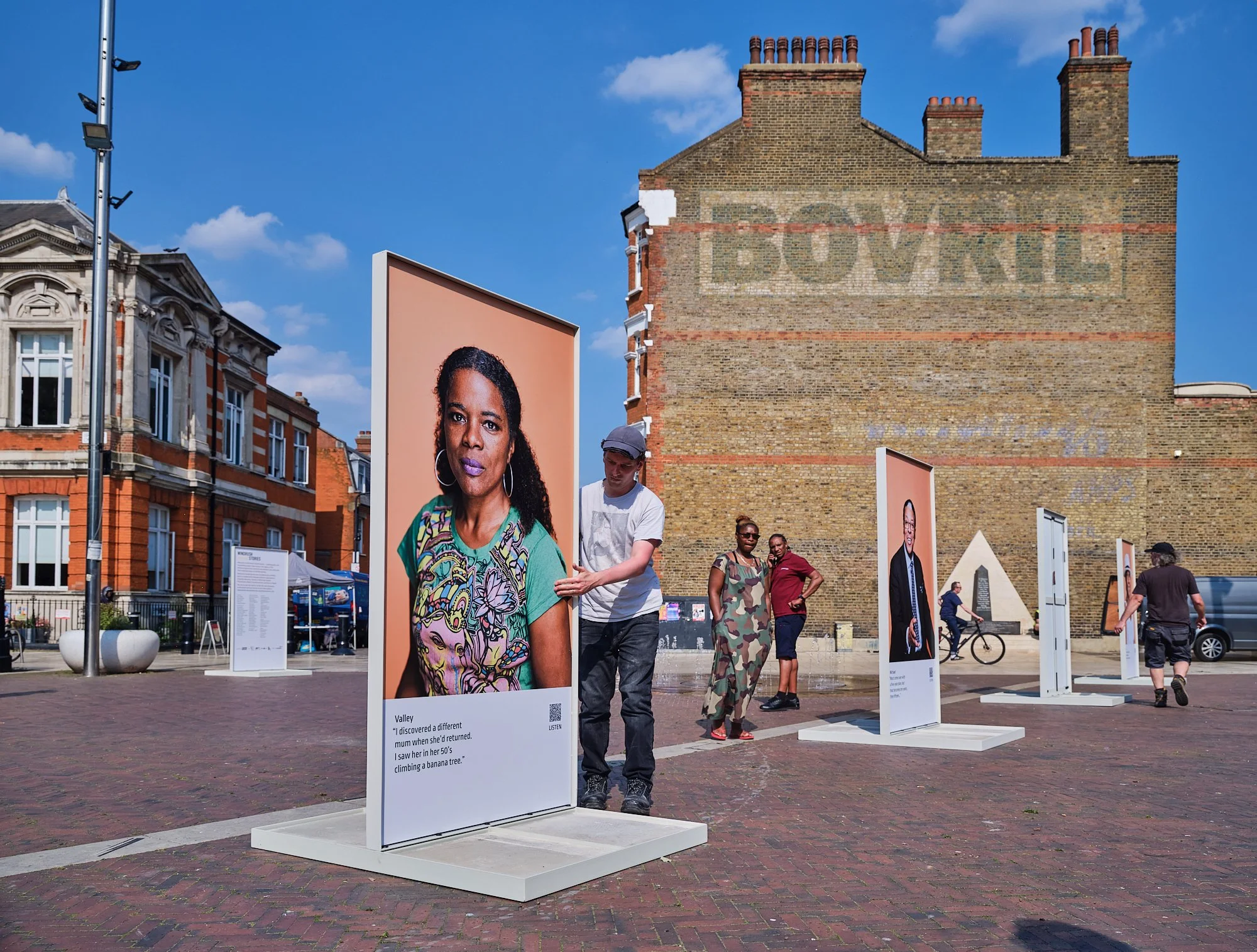 Outdoor art exhibit displaying large portrait photographs on white panels in a city square with historic brick buildings and a clear blue sky.