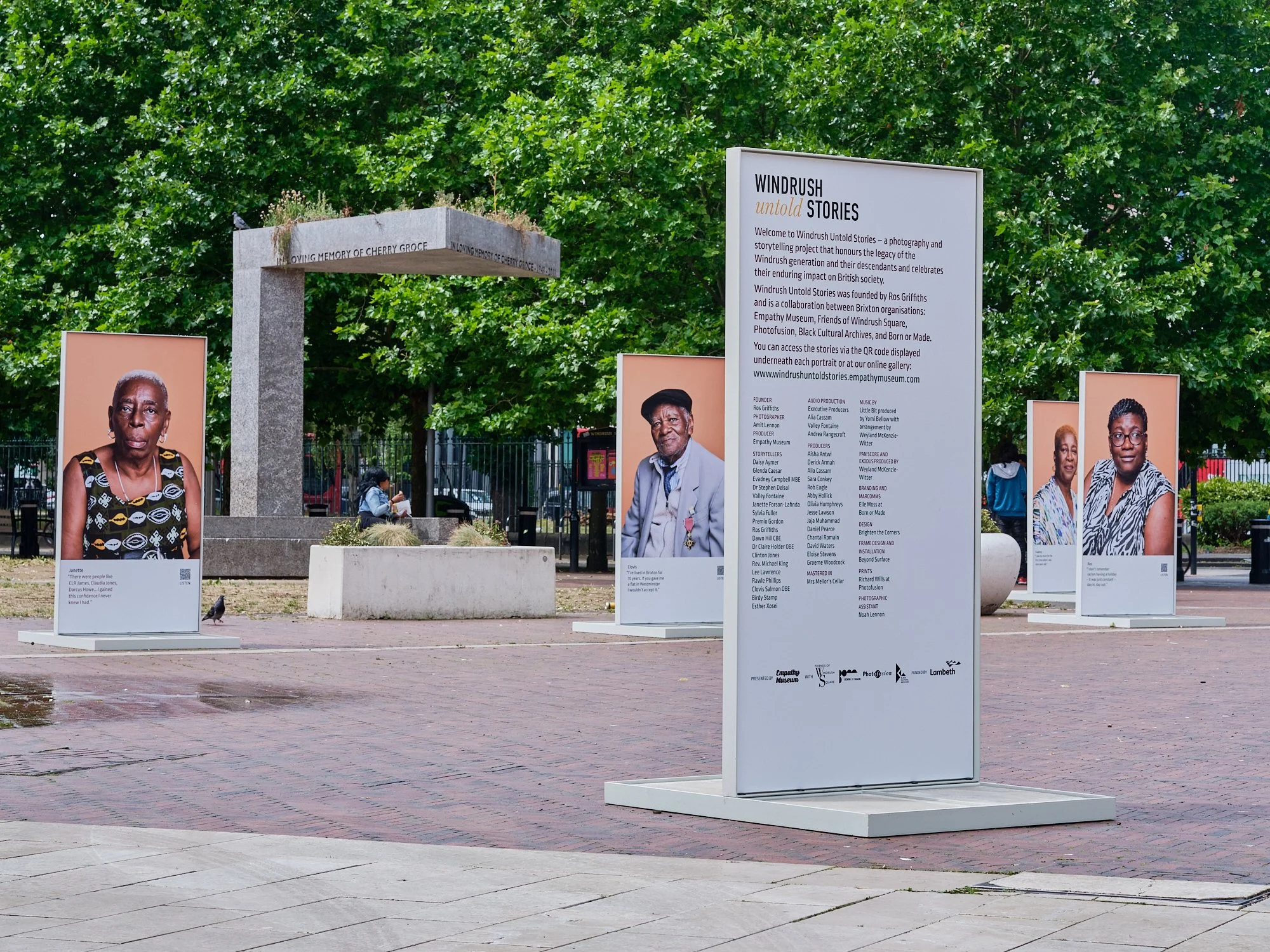 Outdoor public art exhibit with large portrait photos of diverse individuals on white display boards, set in a park area with trees and a concrete structure in the background.