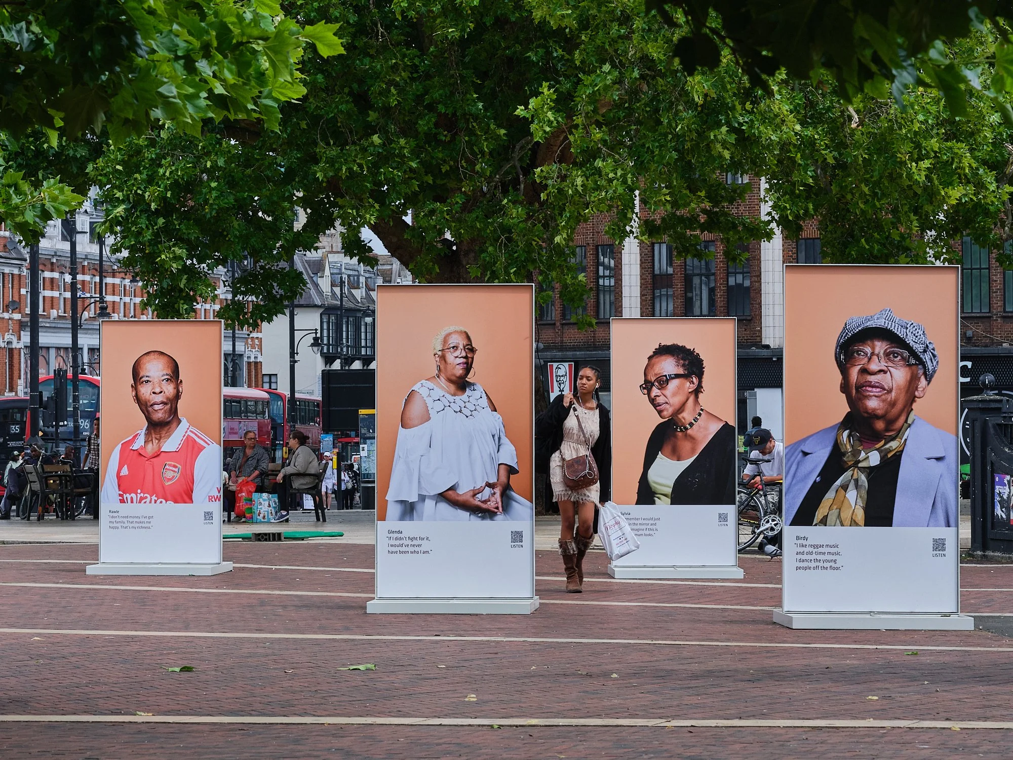 Outdoor exhibition with five large portrait posters on display, featuring diverse older women, set against a city street with trees, brick buildings, and people walking or sitting in the background.