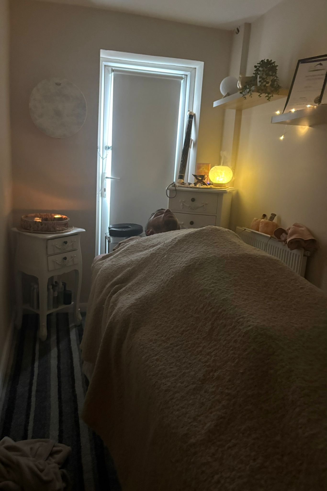 A person lying on their back in a dimly lit room with a bed covered in a beige blanket, a white nightstand with bottles below, a small yellow lamp on a dresser, and wall shelves with decorative items, including a Himalayan salt lamp and a certificate or framed document.