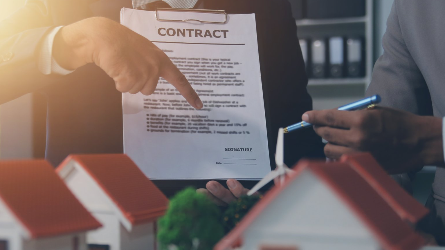 Two people exchanging a contract at a real estate meeting, with model houses in the foreground.