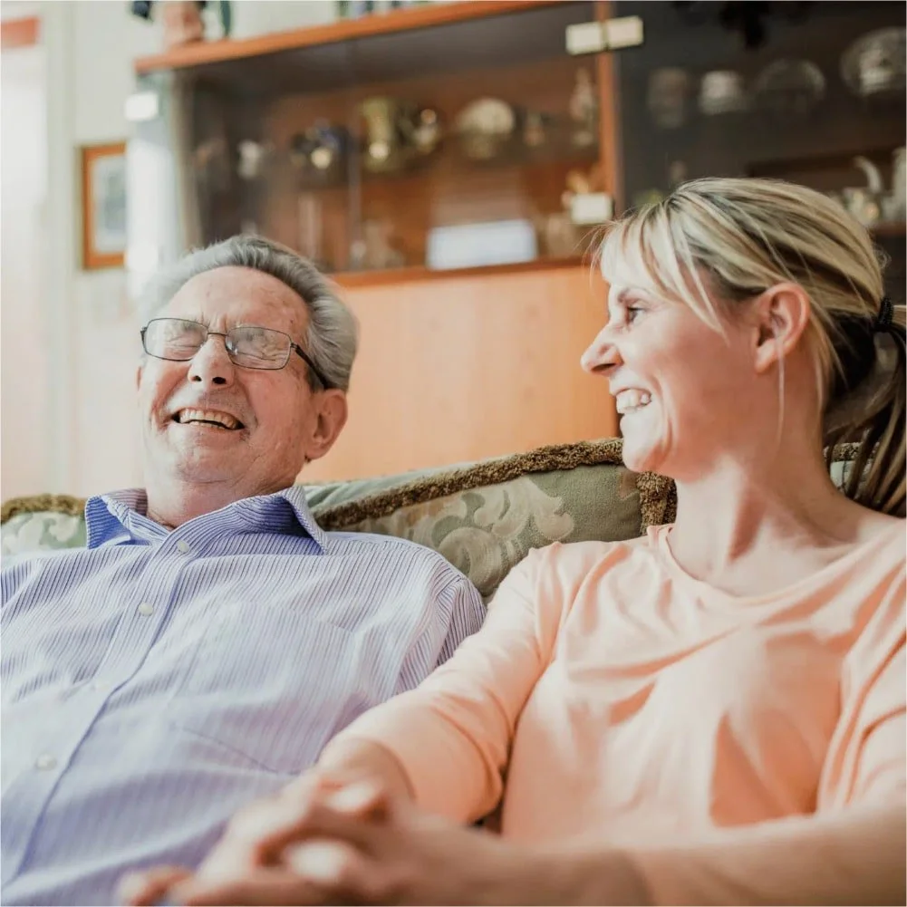 Man with greying hair and round glasses is wearing a striped shirt and listening to a befriender who is wearing a peach long sleeved top and talking to the man
