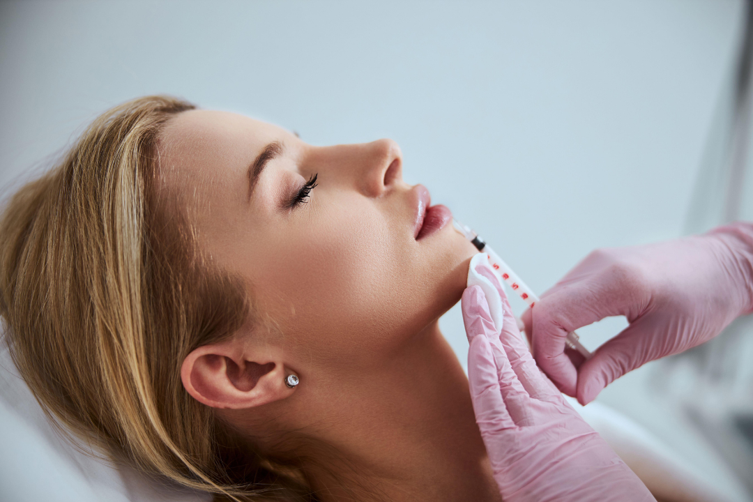A woman lying down with a healthcare professional administering a cosmetic injection to her chin.