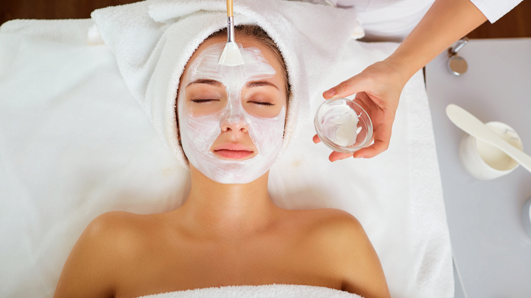 Woman lying on a spa bed with a facial mask being applied, towel wrapped around her head, eyes closed, as a spa worker holds a bowl of facial cream and a brush.