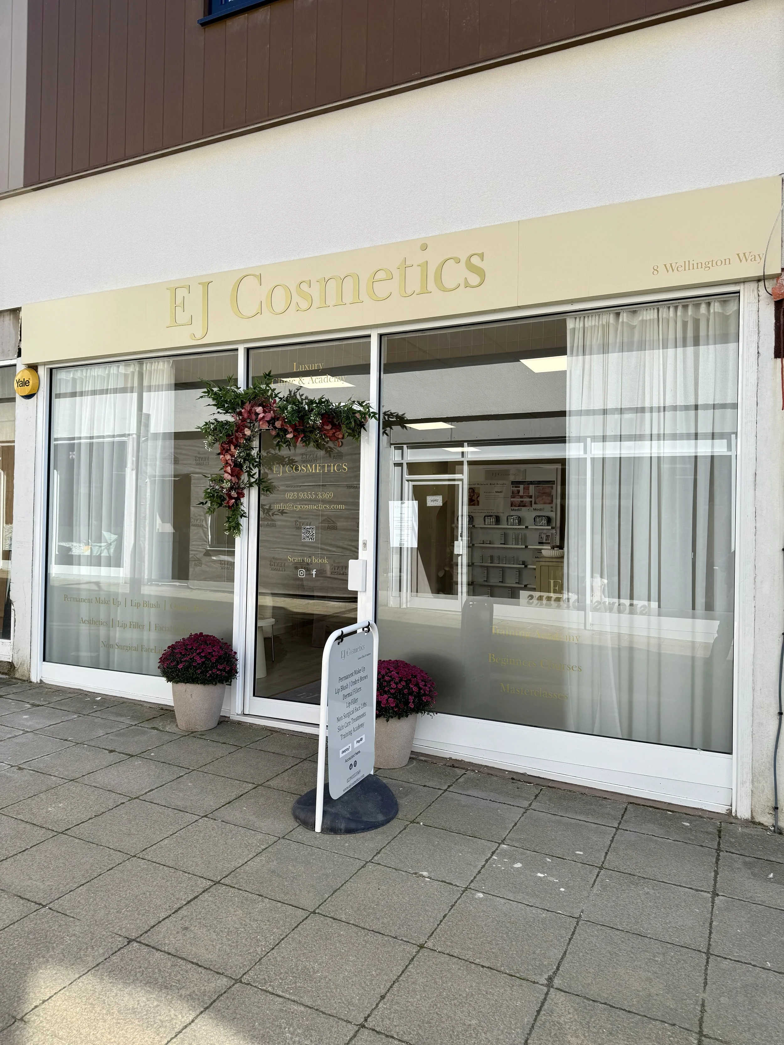 storefront of EJ Cosmetics with gold signage, flowers decorating the entrance, and a signboard outside listing services and contact information, located at 8 Wellington Way.
