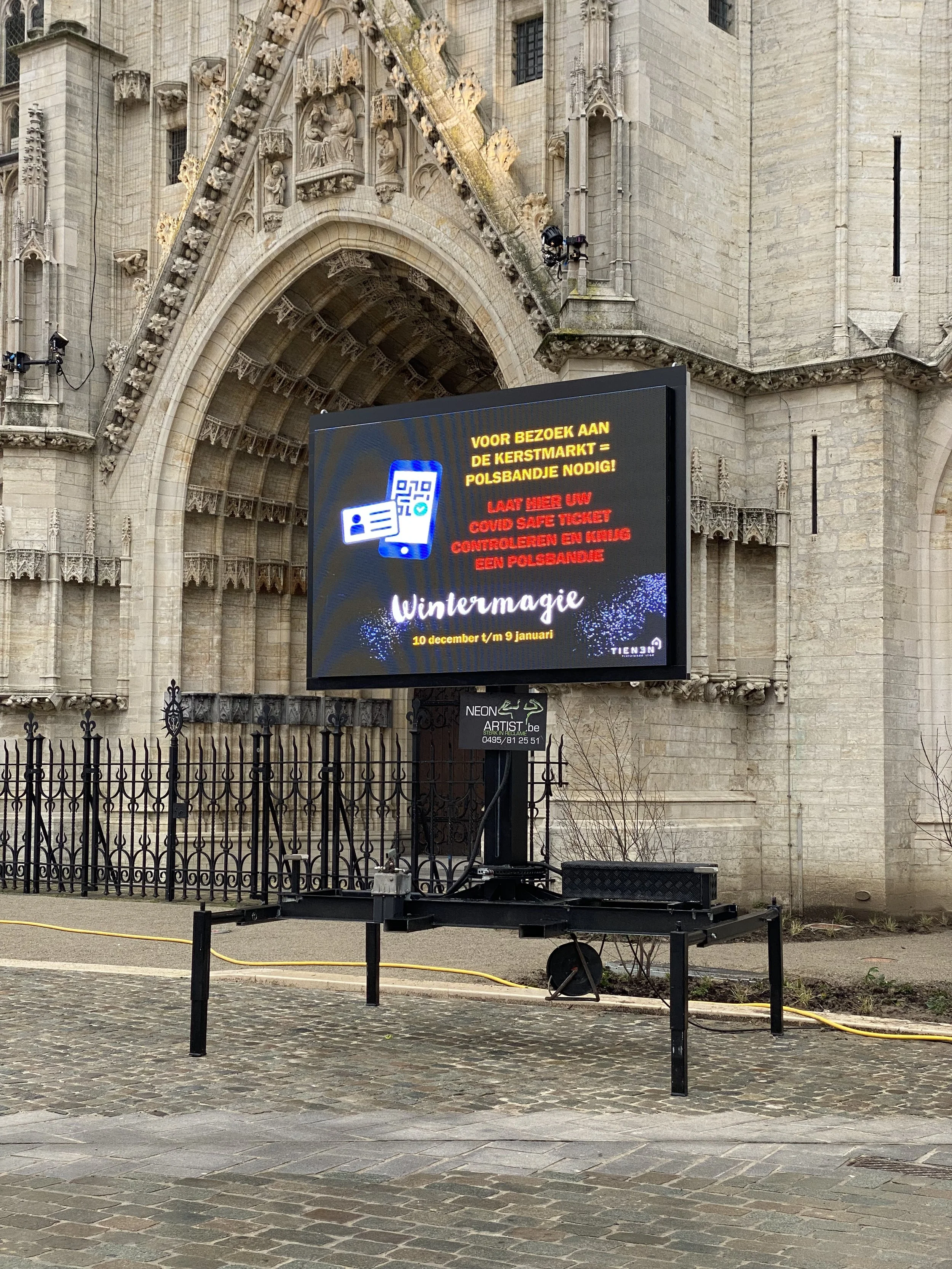 ledscherm buiten, met een sierlijk historisch gebouw op de achtergrond. Het toont een boodschap over COVID-veiligheid op de kerstmarkt en een advertentie voor 'Wintermagie' van 10 december tot 9 januari. Geschikt voor digitale signalisatie