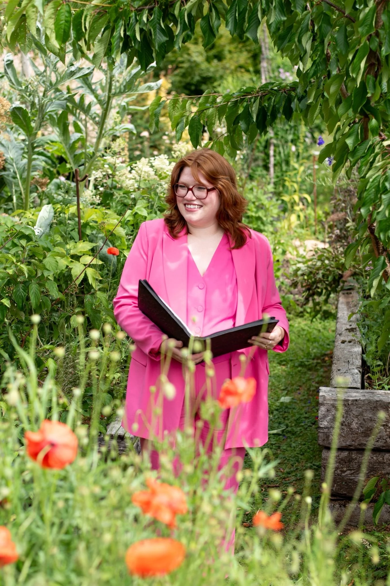 image of annie with ginger hair and a pink 3 piece suit, waiting in a floral garden for the couple to walk down the aisle, she is smiling