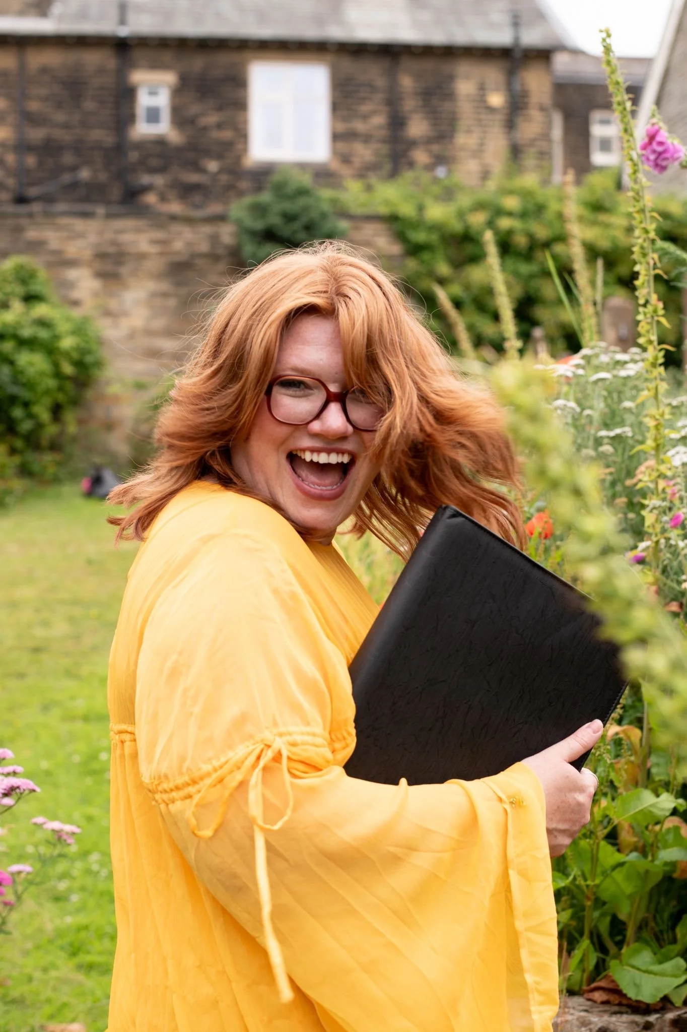 A woman with red hair and glasses smiling joyfully while holding a black folder in a garden with colorful flowers and green shrubs.