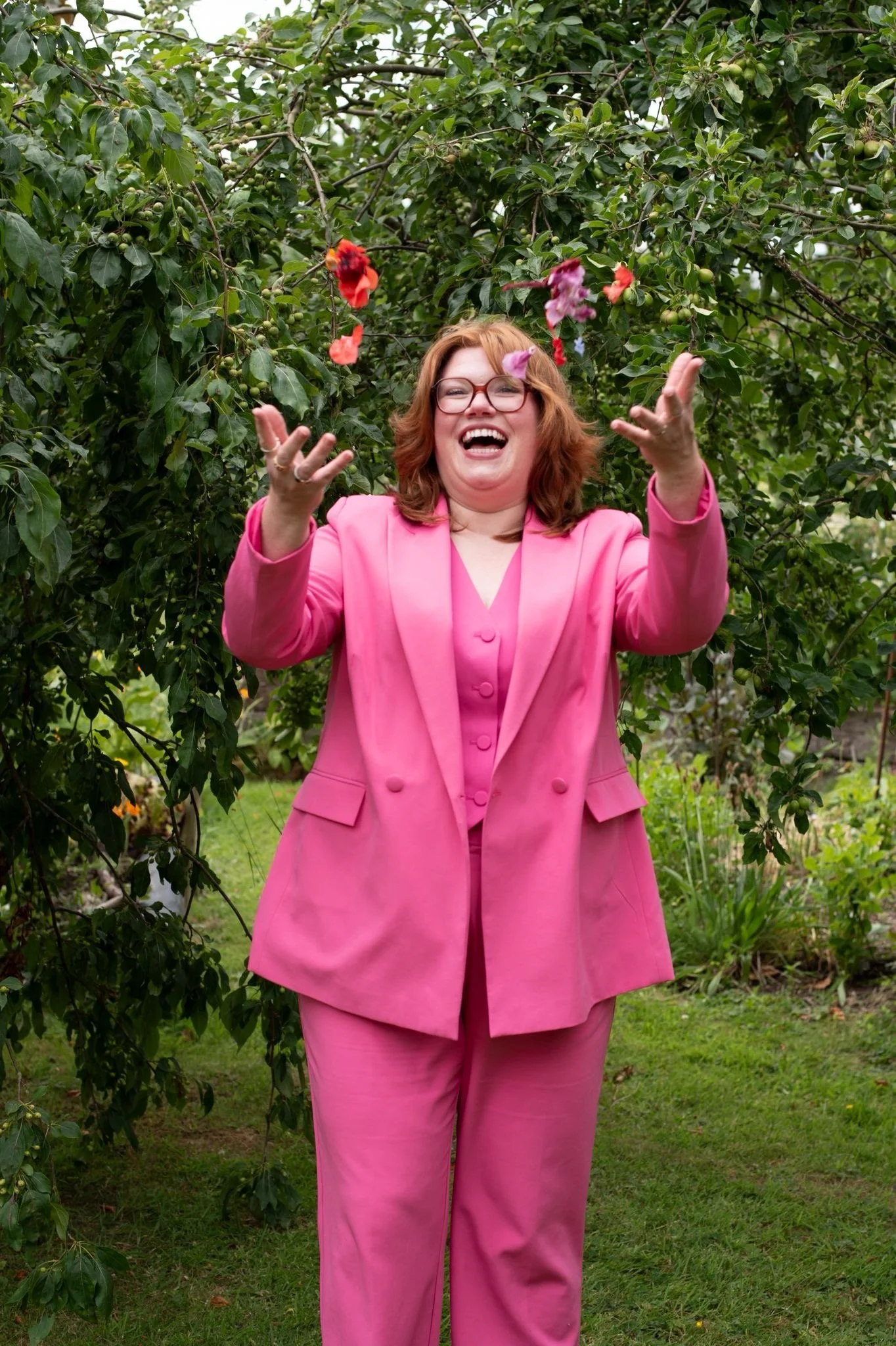 A woman in a pink suit stands outdoors, laughing and throwing flower petals into the air.