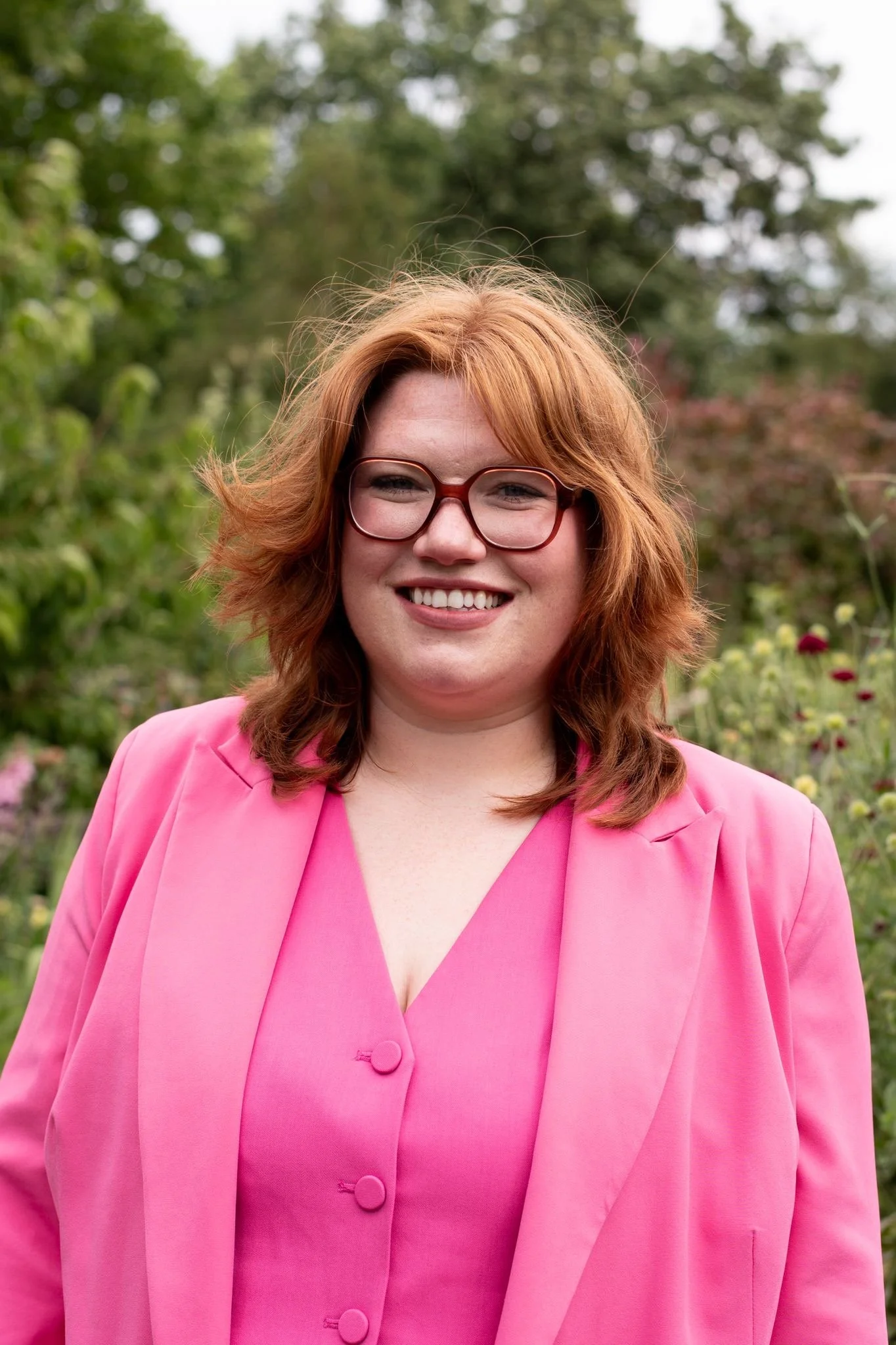 A smiling woman with red hair, glasses, wearing a pink blazer and dress, standing outdoors with green trees and flowers in the background.