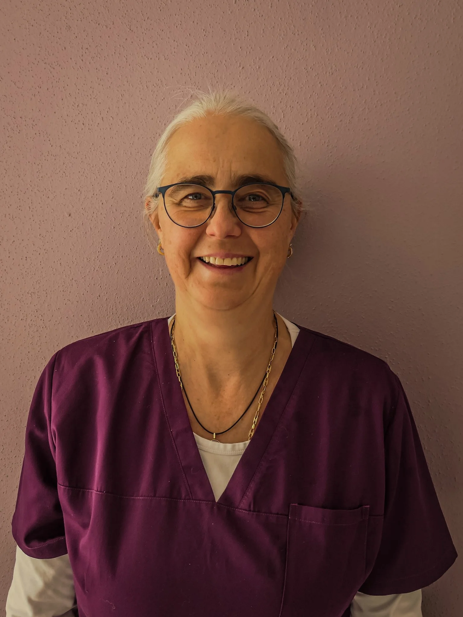 Eine lächelnde ältere Frau mit weißen Haaren, Brille, in einer medizinischen dunkelvioletten Uniform vor einer pinken Wand.