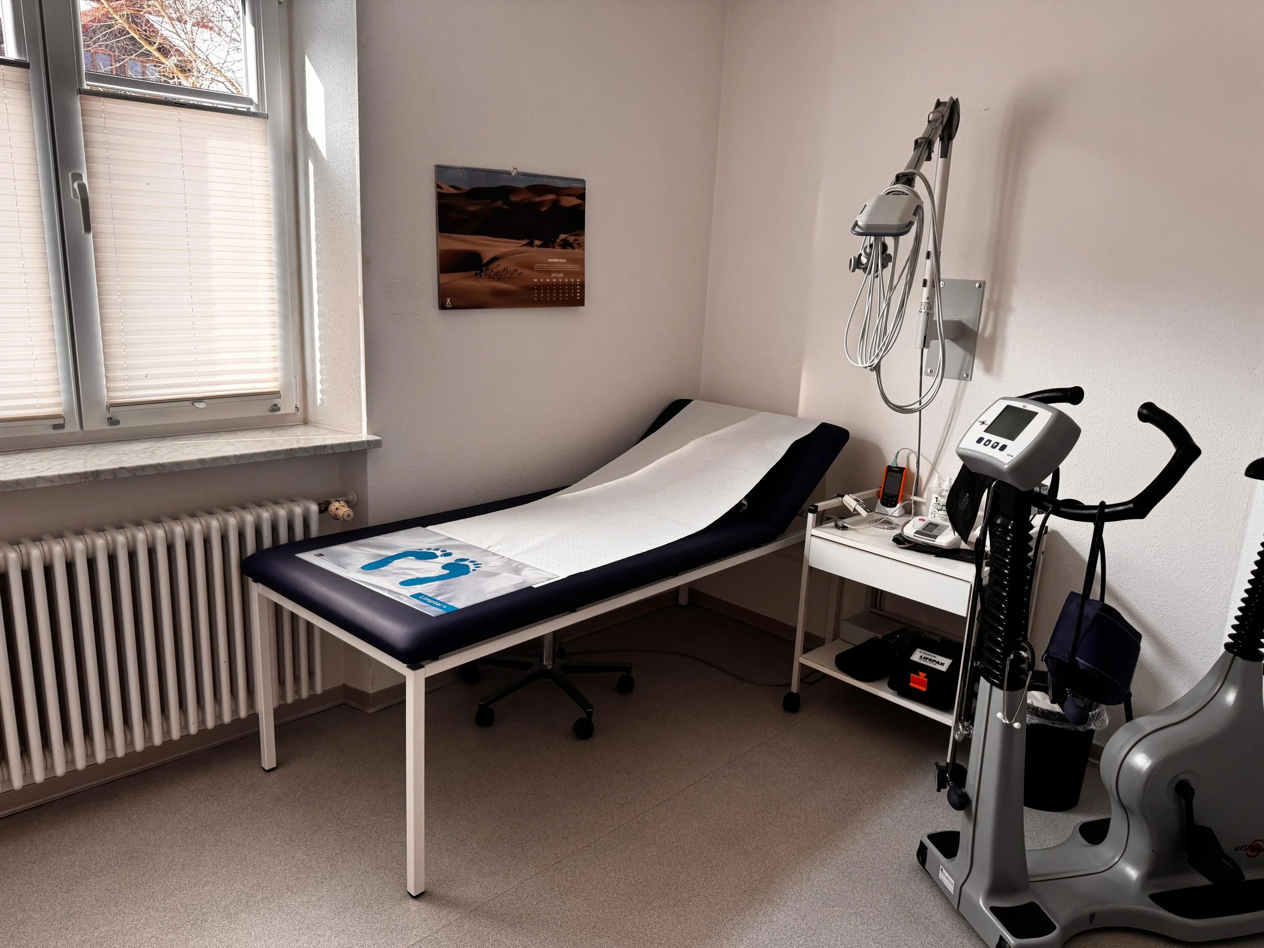 Ein Arztpraxisraum mit Untersuchungsliege, medizinischen Geräten und einem Fenster mit Rollos.