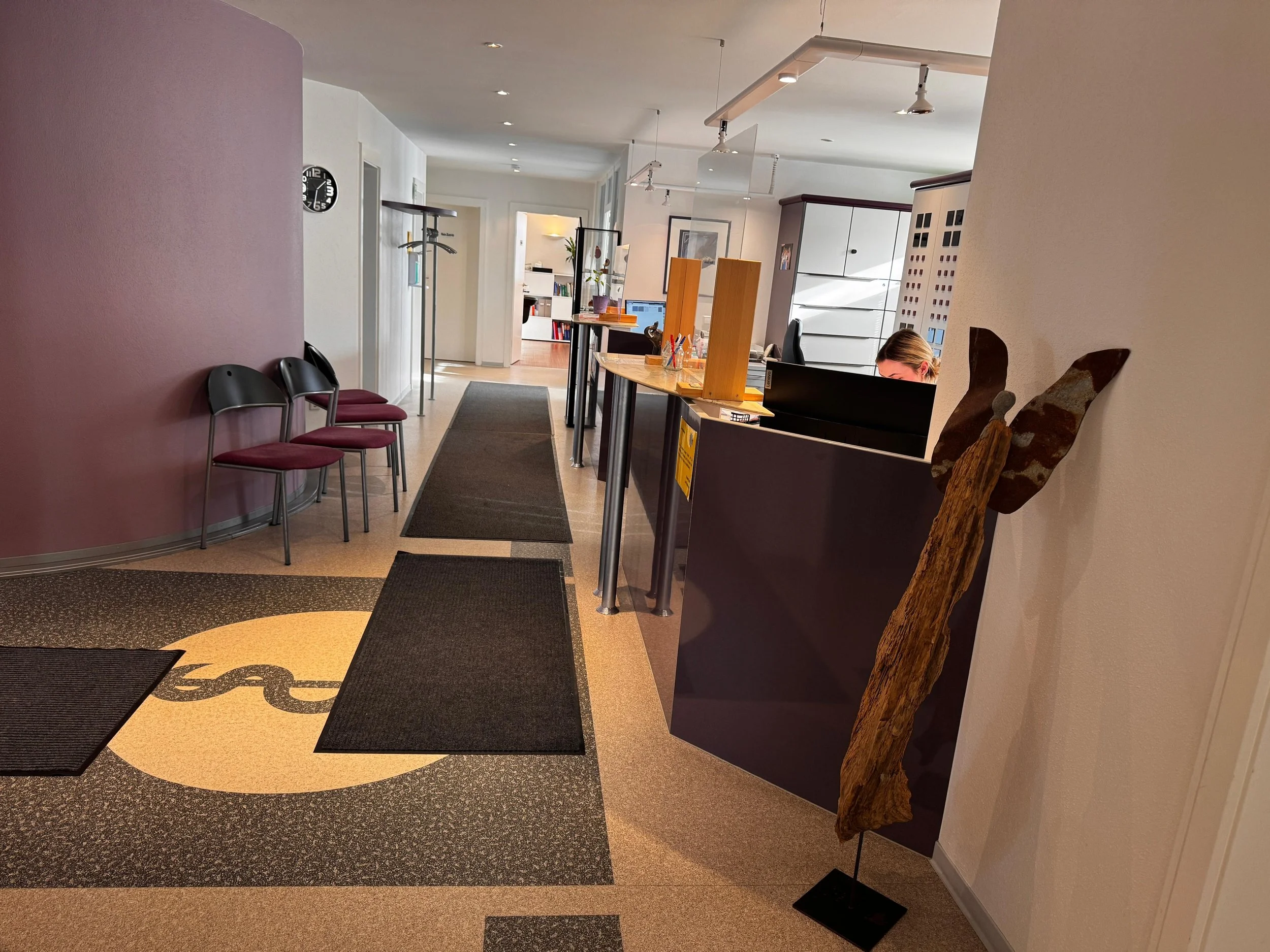 Rezeption eines Wartebereichs mit drei schwarzen Stühlen mit roten Sitzkissen, zwei schwarzen Teppichen, einer Holzskulptur neben der Rezeption, einem Empfangstresen mit einem Computer und einer Mitarbeiterin, weiteren Möbeln und einem Flur, der in einen Raum führt.